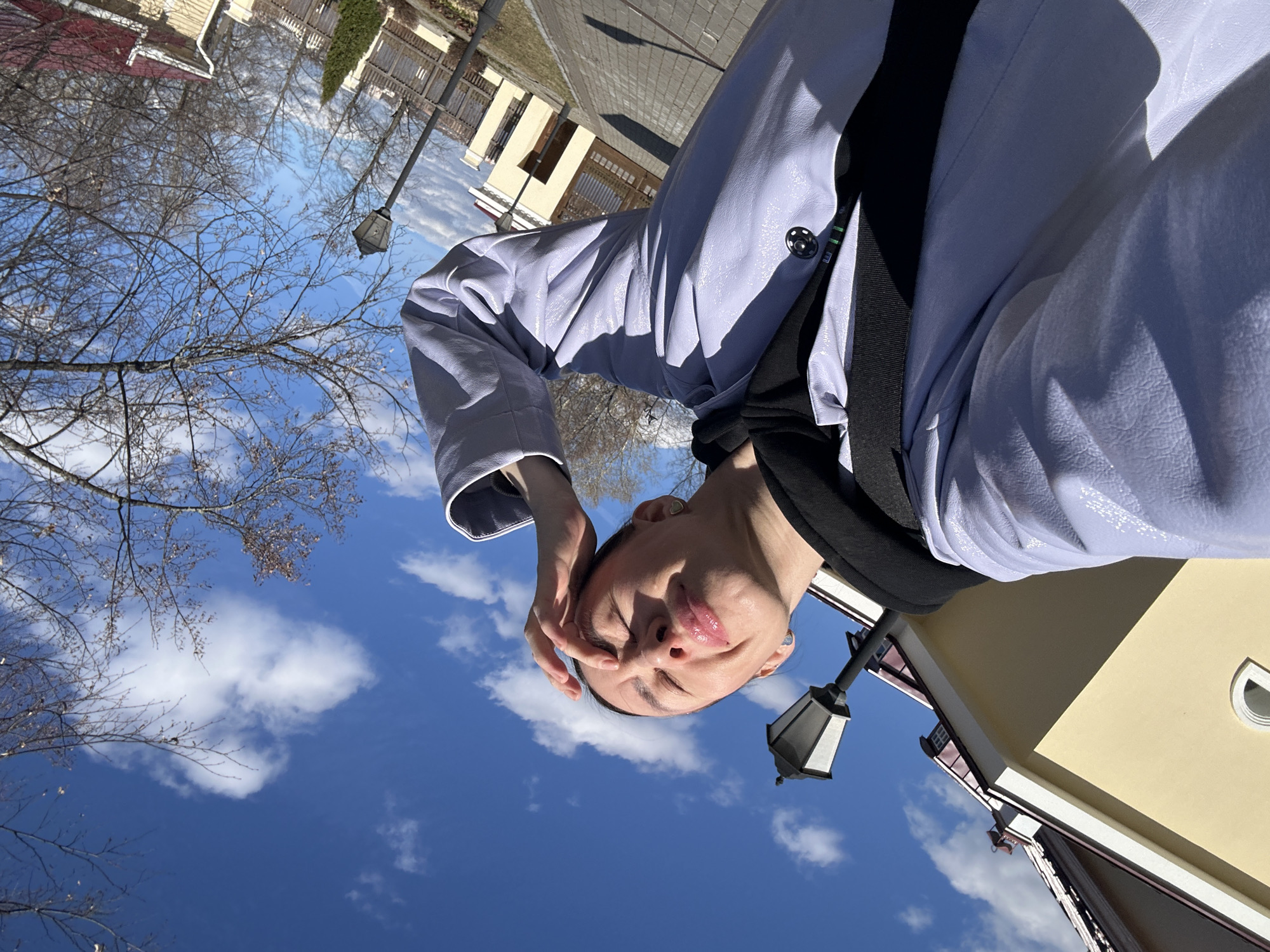 A person is taking a selfie outside in Votkinsk, Russia. The camera angle is tilted, with the person's face in the foreground and the sky in the background. The person is wearing a light-colored jacket with a black strap. A street lamp is visible to the right of the person's head. Behind the person, there are buildings and bare trees against a blue sky with scattered clouds. The ground appears to be paved.