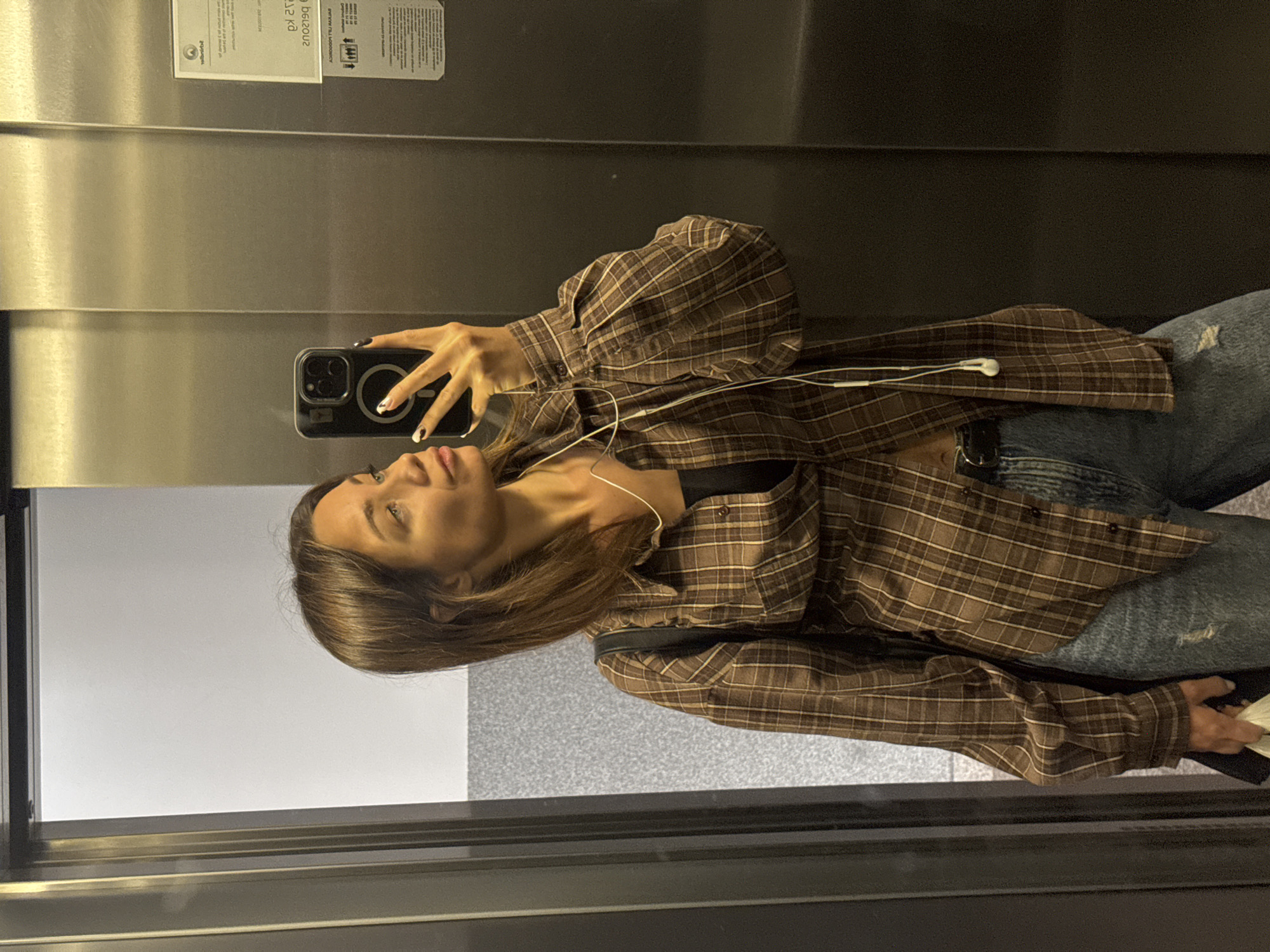 The image is an eye-level shot of a woman taking a selfie in what appears to be an elevator. She is wearing a brown plaid shirt over a black top, blue jeans and white earphones. She is holding a black cell phone. The background is a metal elevator and glass with a view. The exact location in Budva, Montenegro, is not identifiable from the image.