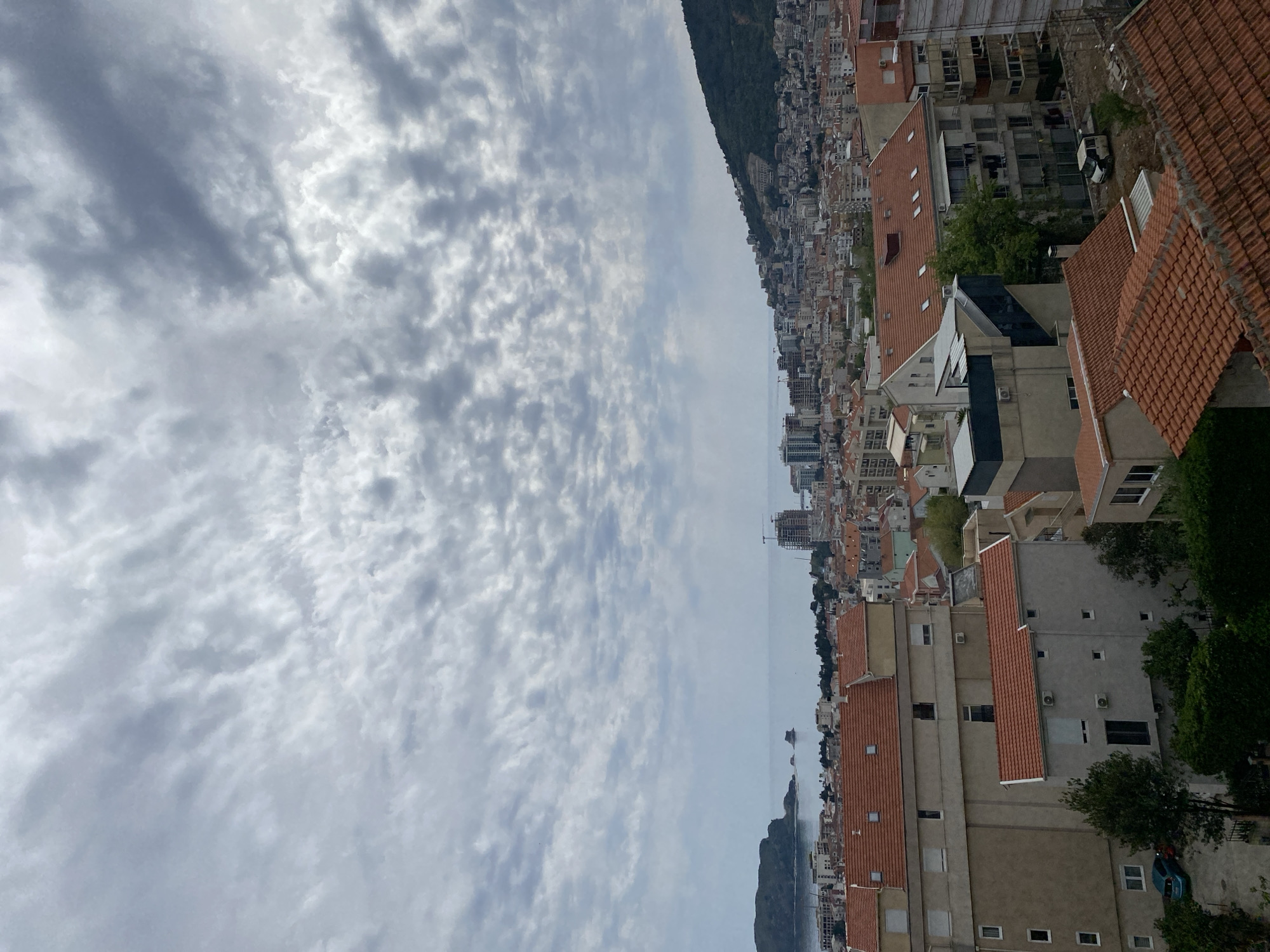 The image captures a sideways view of the city of Budva, Montenegro, under a cloudy sky. Buildings with red-tiled roofs dominate the right side of the frame. A mountain ridge is visible in the distance. The Adriatic Sea is discernible between the buildings and the mountain. No people are directly visible.