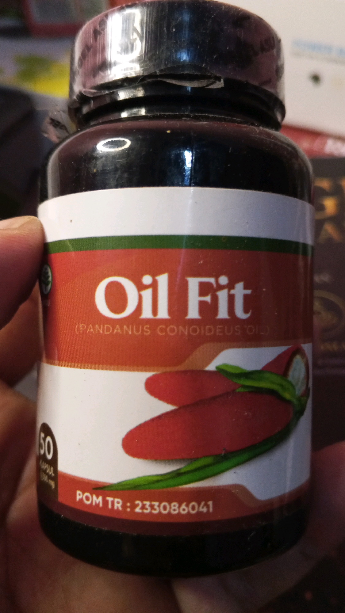 The image shows a close-up of a bottle of "Oil Fit" supplements. The bottle has a black cap and a label that is predominantly white and orange. The label prominently displays "Oil Fit" in large white letters. Below this, in smaller white text, it reads "(PANDANUS CONOIDEUS OIL)". To the left of this text, there is a small green circular logo. On the lower part of the label, there is a section with red and green imagery, appearing to be stylized depictions of peppers. To the left of this imagery, there is a circular area with the number "50" in large white font, followed by smaller text that is difficult to read but appears to say "KAPSUL". At the very bottom of the label, there is a red band with white text that reads "POM TR : 233086041". The bottle is held by a person's hand, with fingers visible on the left side of the image. The background is blurred but suggests an indoor setting. There is no indication of the city of Semarang, Indonesia, in the image itself.