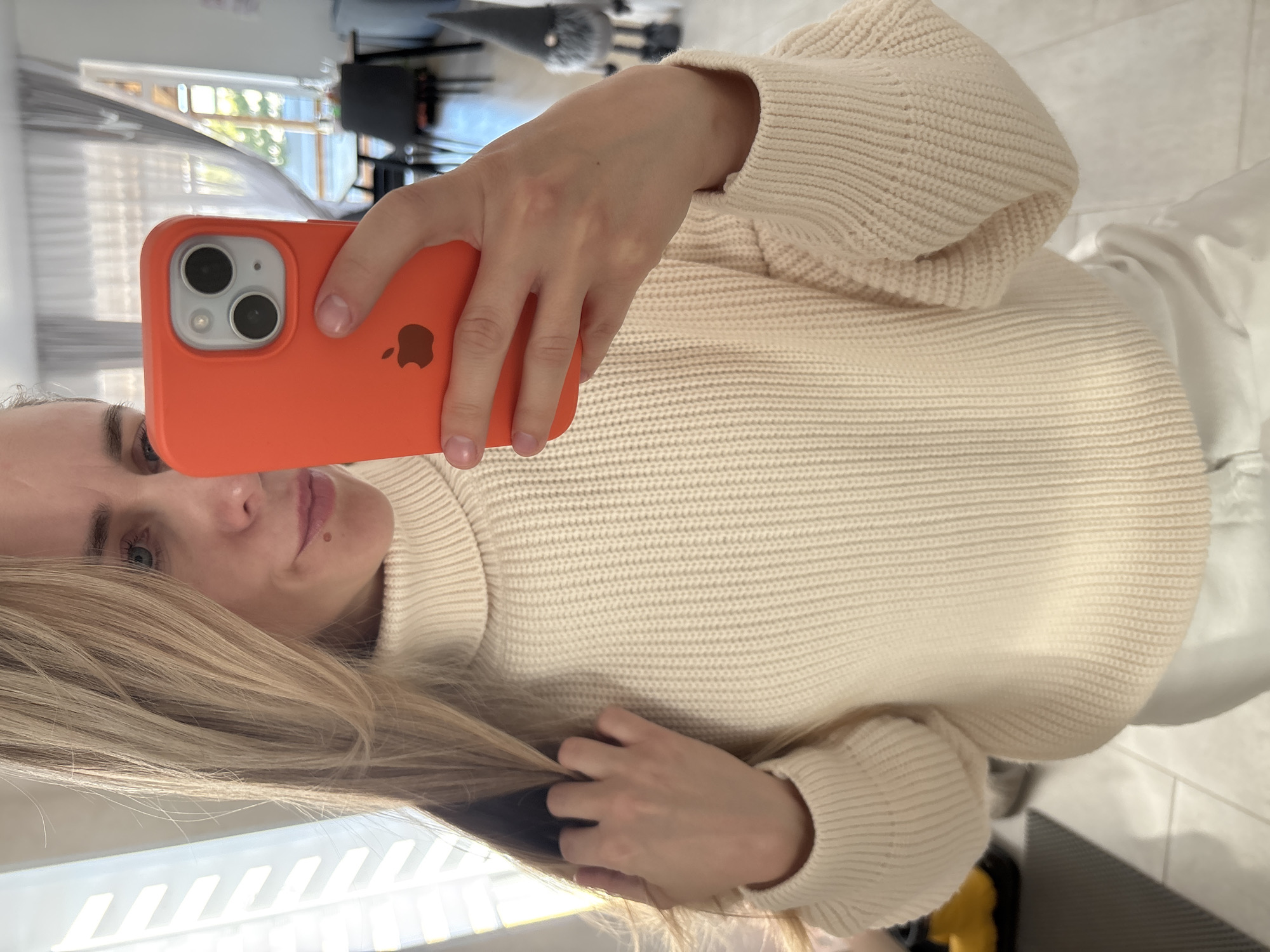 The image, oriented sideways, captures a mirror selfie of a woman in an indoor setting, contextually located in Chaikovsky, Russia. The woman has light skin, long blonde hair that partially covers the left side of her face, and visible blue eyes. She has a neutral expression with slightly parted lips and a small mole above her upper lip.
She is wearing a cream or off-white chunky knit turtleneck sweater with ribbed cuffs and hem. Her right hand, with natural, short nails, holds an orange iPhone in a matching case, displaying the brown Apple logo on its back. The phone's dual camera lenses are clearly visible as it captures the reflection.
The background, seen through the mirror reflection, shows a window letting in natural daylight, suggesting it is daytime. Horizontal blinds or curtains are visible in the window. Reflections of dark-colored furniture, possibly chairs or a desk, can be seen behind the window. The floor visible in the reflection appears to be light-colored tiles. The woman's left hand is gently touching her hair near her shoulder.