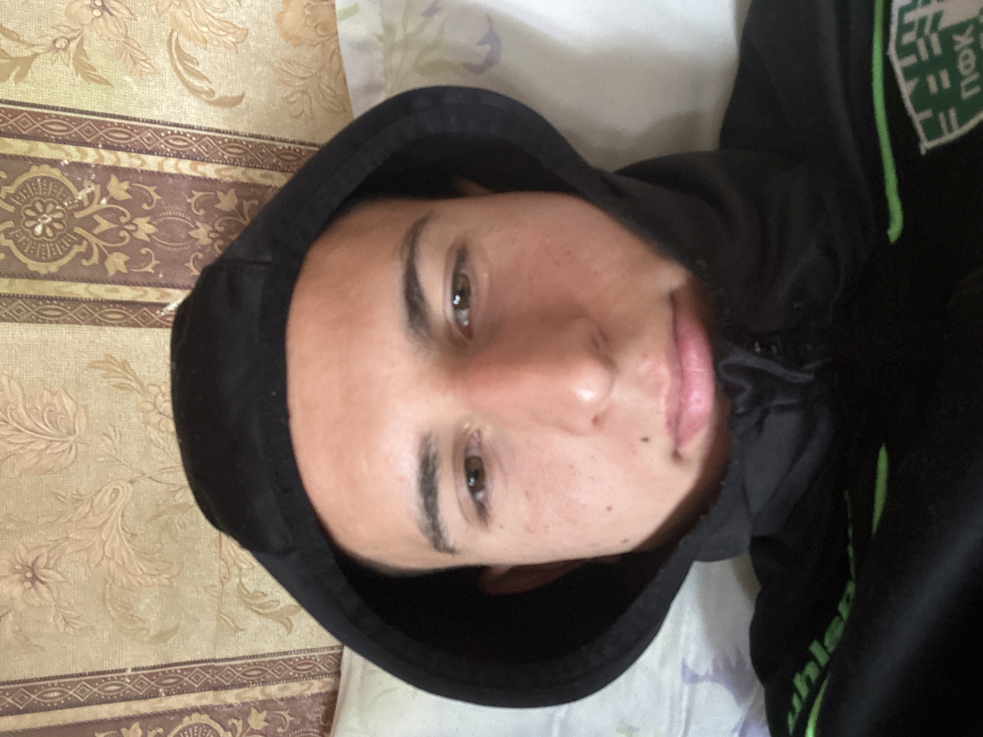 A person wearing a black hooded sweatshirt with green lettering is lying on a bed with a patterned bedspread. The image appears to be taken indoors. No information is available to corroborate location details.