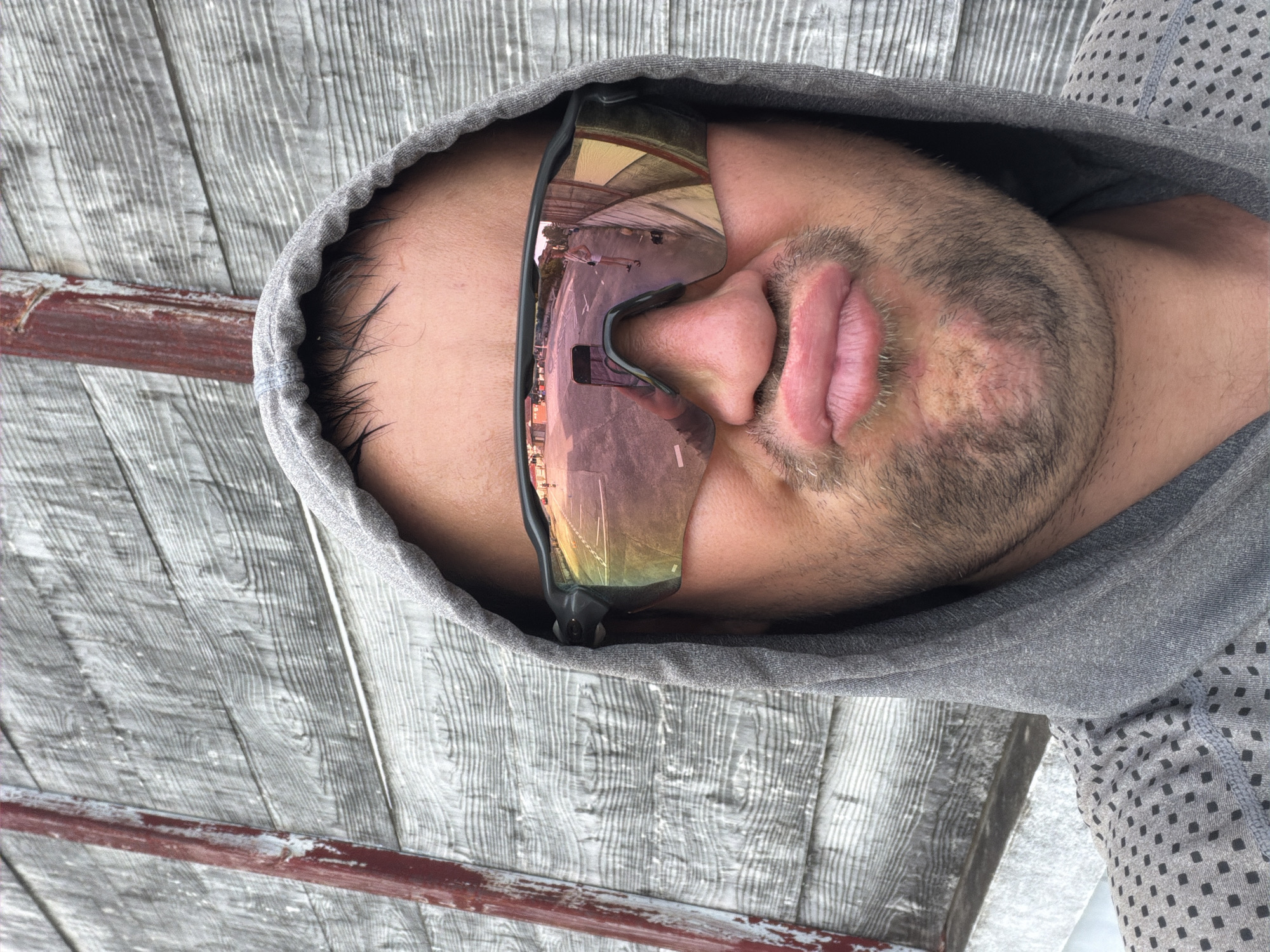 A close-up, low-angle selfie shows a Caucasian man in a grey hooded sweatshirt and reflective sunglasses, puckering his lips. He stands in front of a weathered, grey wooden wall with a vertical red beam on the left side. The sunglasses reflect a street scene with buildings, a road, and a person standing. The man has dark hair, stubble, and fair skin. The image appears to be taken outdoors. It's not possible to determine the exact location in Baan Mae Nam, Thailand from the image alone.