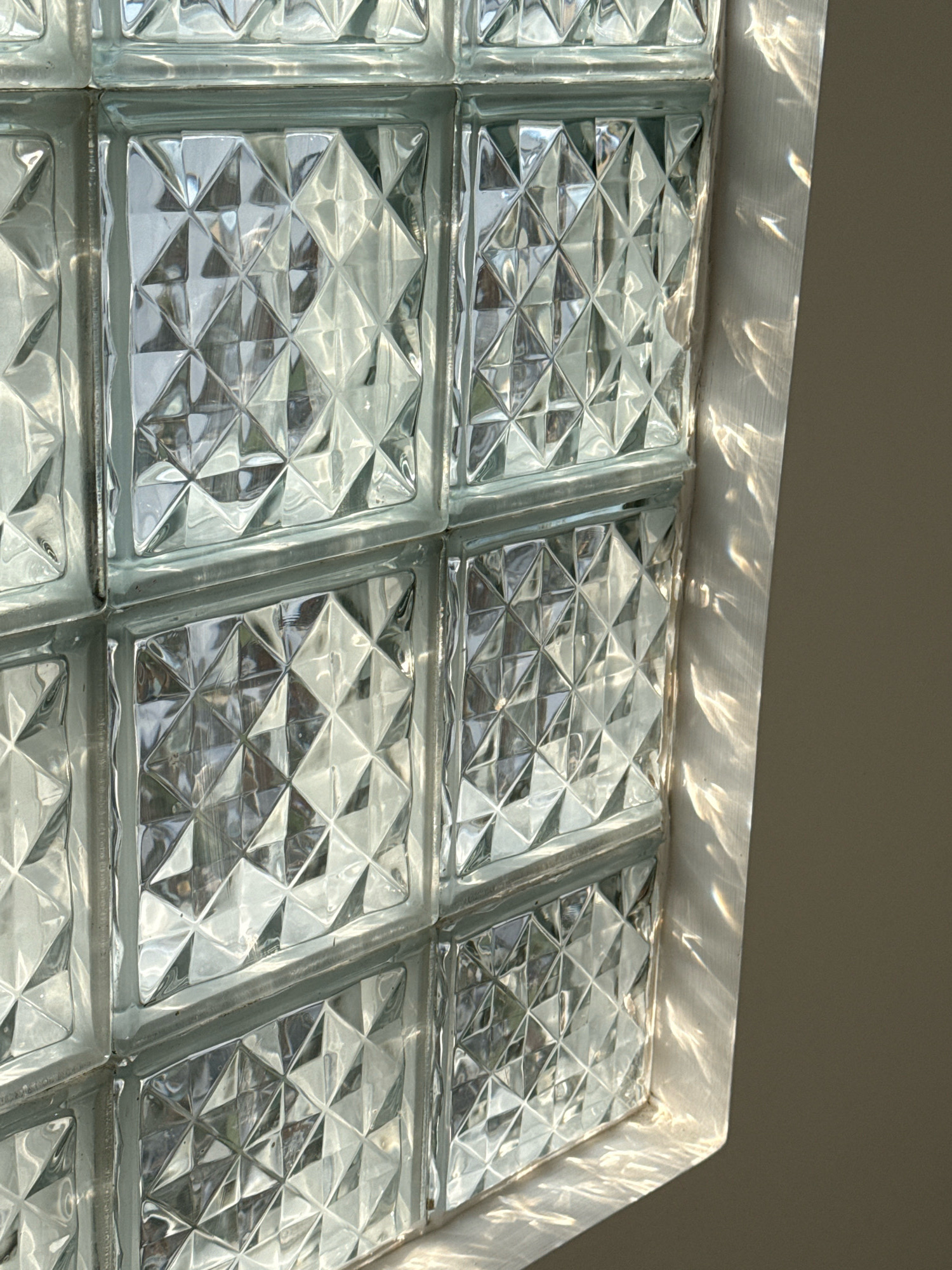 The image displays a section of a glass block wall, likely in an interior setting in Nha Trang, Vietnam. The glass blocks have a textured, diamond-shaped pattern on their surface, which refracts and reflects light. Sunlight is casting bright, shimmering patterns onto a beige wall adjacent to the glass block wall. The glass blocks appear to be arranged in a grid pattern, with visible grout lines between them. The lighting suggests it is daytime, with the sun positioned to create these light effects. There are no people or explicit actions depicted. The scene is clean and devoid of clutter, focusing solely on the visual interplay of light and the glass block texture.