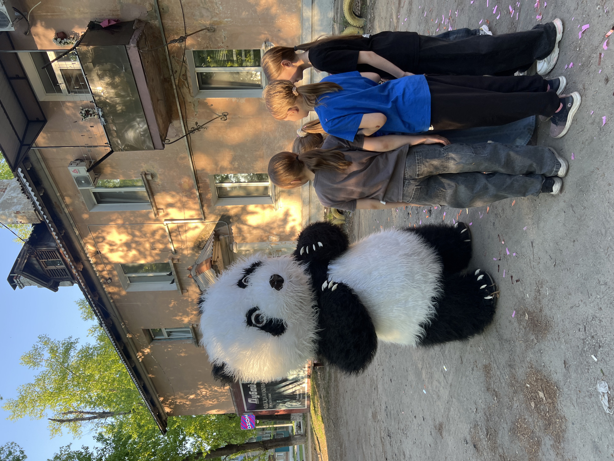 The image depicts three people and a large panda mascot lying on a paved surface in front of a building. The individuals, who appear to be children, are standing in a line, their bodies aligned perpendicularly to the mascot, who lies on its back. The building behind them has multiple windows and what appears to be an older facade. A tree and a signpost are visible to the left of the building, suggesting an urban or suburban street location. The ground has scattered confetti. The city and region are Unknown.