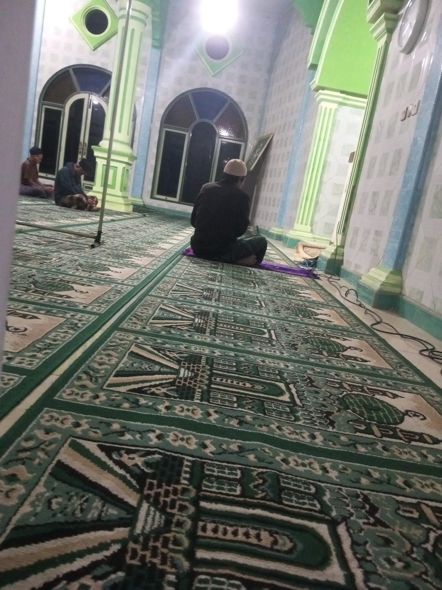 The image depicts the interior of a mosque in Andongsili, Indonesia, featuring a large, intricately patterned carpet that covers the floor. Several individuals are present, all dressed in traditional Islamic attire, engaged in prayer. Two men are seated on the carpet in the foreground, facing away from the camera, while another man is seated closer to the wall on the right side of the frame, also in prayer. The walls of the mosque are decorated with tiled patterns and feature arched windows. Green and white columns are prominently visible, supporting the ceiling. A small clock is mounted on the wall in the upper right corner. The lighting appears to be artificial, casting a soft glow throughout the space.