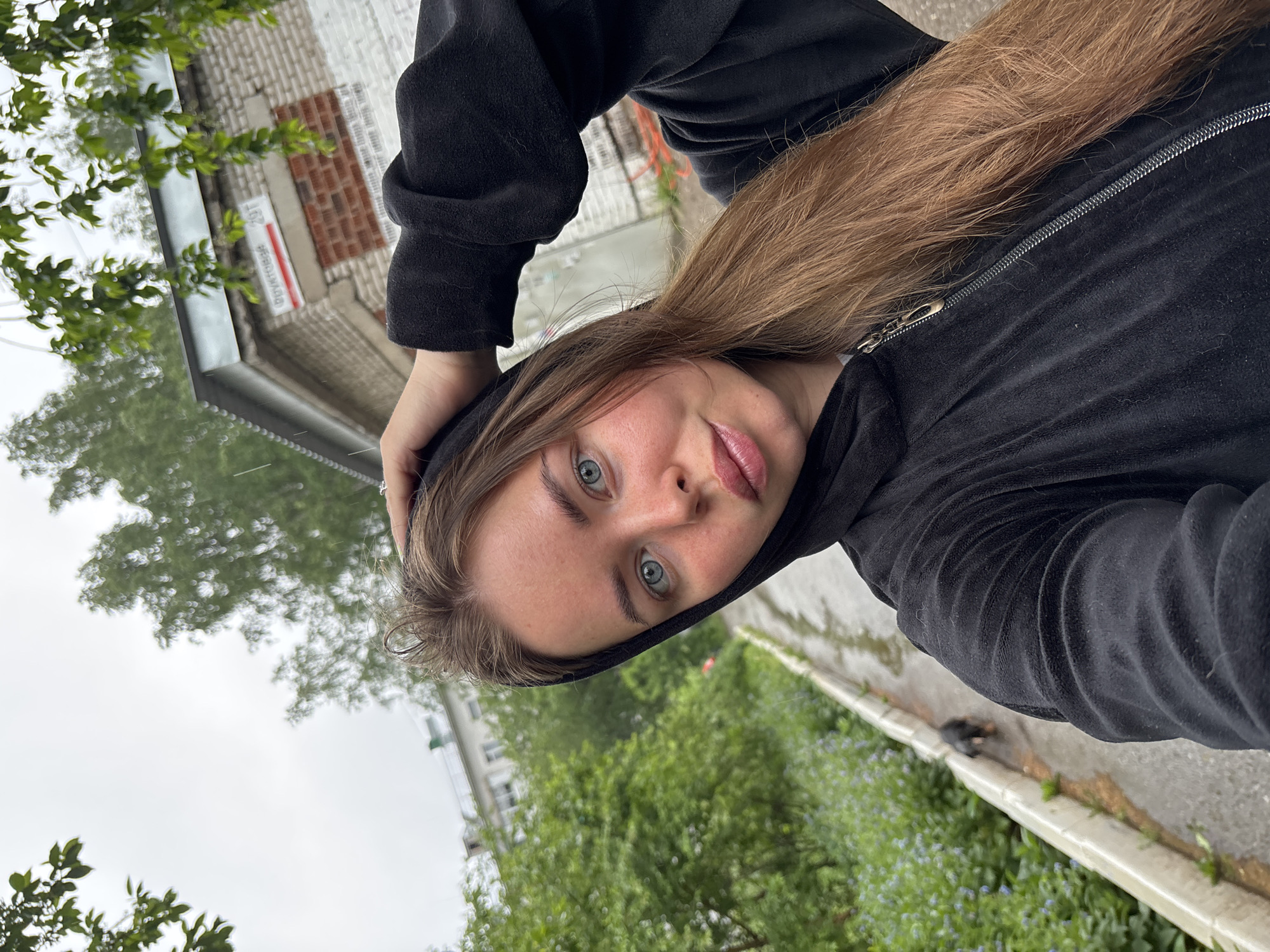 A young woman is wearing a black velour hoodie with the hood up. She is looking directly into the camera in a selfie. The location is Izhevsk, Russia, as indicated by the street sign visible in the background, which reads "157 Berkhgolts." She is standing near a curb with vegetation. The sky is overcast, and it appears to be raining.