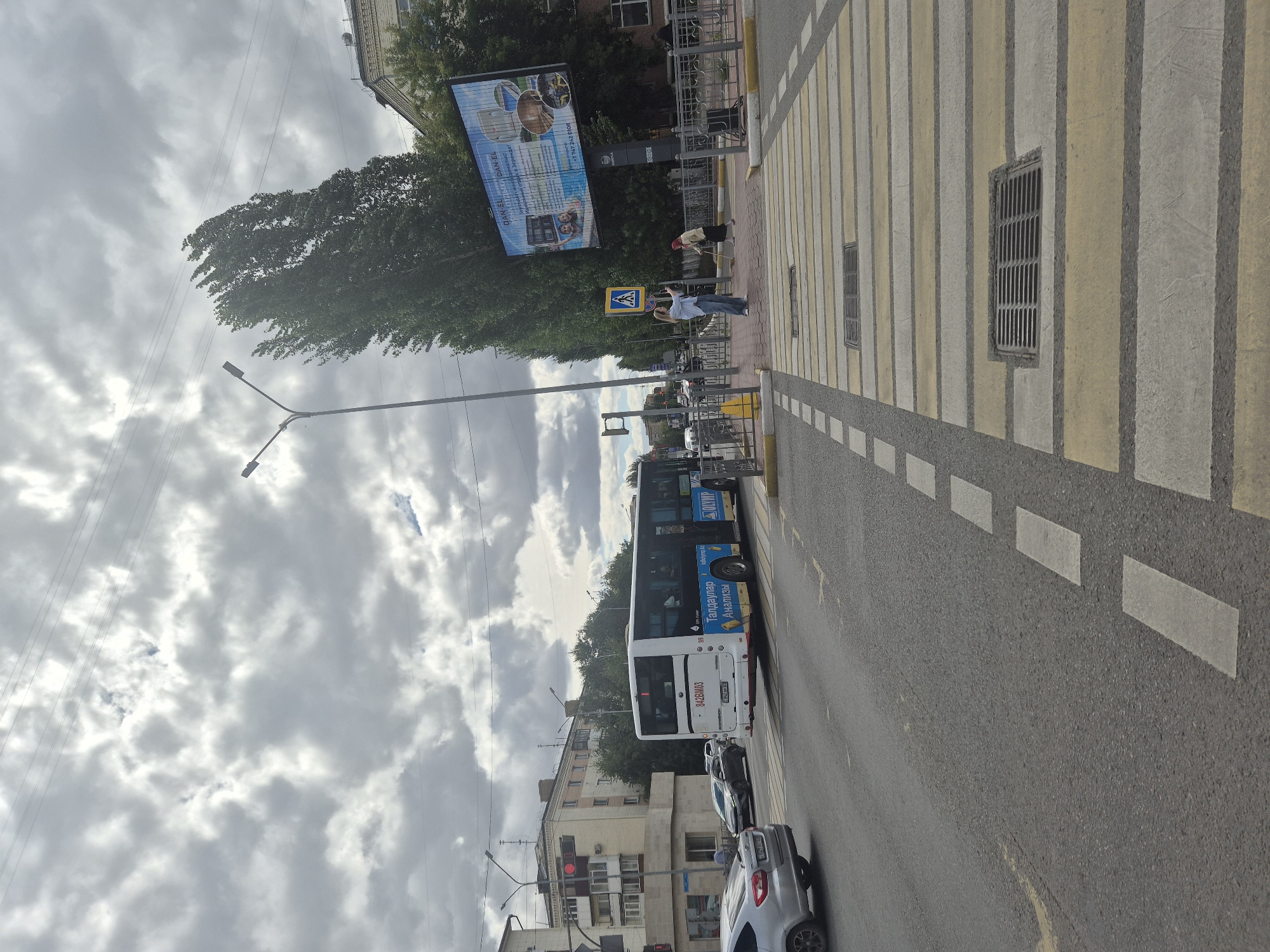 A wide-angle, eye-level, outdoor shot captures a street scene in Kokshetau, Kazakhstan. A white bus with blue advertisements is turning right at a pedestrian crossing. The bus has the license plate 842BMO3. On the sidewalk to the right, two women are present. One, in a light blue shirt and jeans, holds up a phone, possibly taking a picture. The other, in a yellow headscarf and dark clothing, is holding a cane and walking. A large billboard with advertisements is visible behind them, featuring images of buildings and people. The sky is overcast with heavy, gray clouds. The street has a pedestrian crossing with yellow and white stripes. Various cars are visible on the road, including a silver car in the foreground. Buildings and trees line the street. A traffic light is red on the left side of the image.