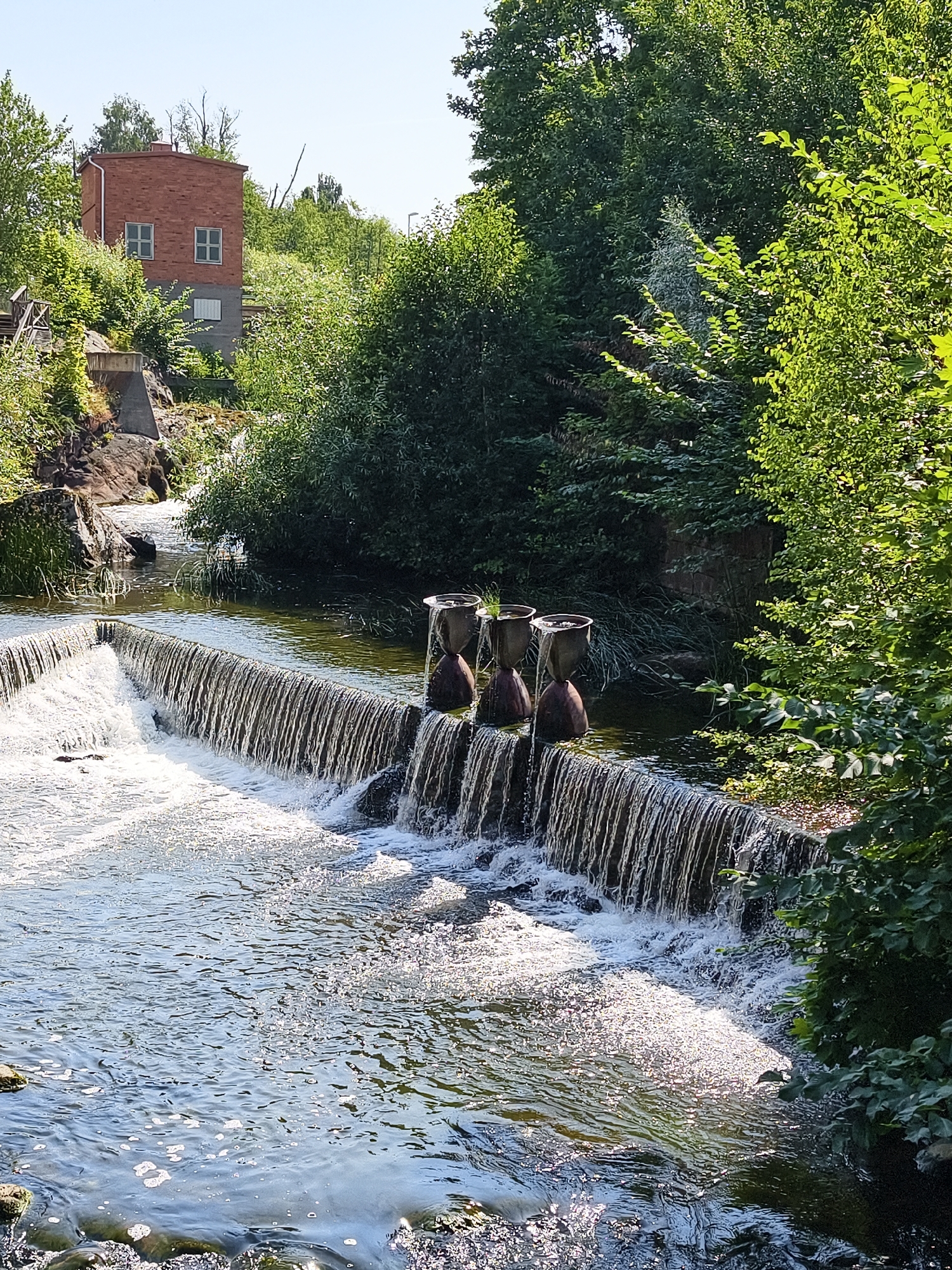 The image depicts a waterfall in a natural setting, possibly part of a park or recreation area. In the foreground, a dam across a body of water creates a cascading waterfall. Several decorative urn-like objects are placed on the edge of the dam. The water is flowing rapidly, creating white foam and ripples. Lush green trees and foliage surround the waterfall, with a clear blue sky overhead. In the background, a red brick building with windows is visible on a hillside, partially obscured by trees. This location is the Kinda Canal waterfall in Linköping, Sweden. There are no people visible in the image.