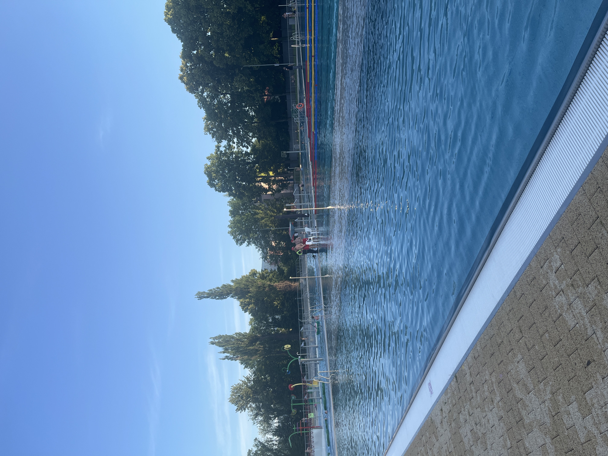 The image shows a large outdoor swimming pool on a clear, sunny day. The pool is filled with blue water, and there are lane markers in red, yellow, and blue in the far section of the pool. The pool area is paved with light-colored bricks and features a white border with a ribbed drainage system.
In the background, there are several tall, slender trees and some buildings. Scattered around the pool, there are people in various activities. In the shallow section of the pool, near the left side, there are several water features, including sprinklers and colorful arches. A group of people, some wearing red shirts and hats that suggest they might be lifeguards, are standing near the edge of the pool. One person is sitting in a white chair. Other individuals are visible swimming or walking in the pool area. The sky is a clear, bright blue with a few wispy clouds.