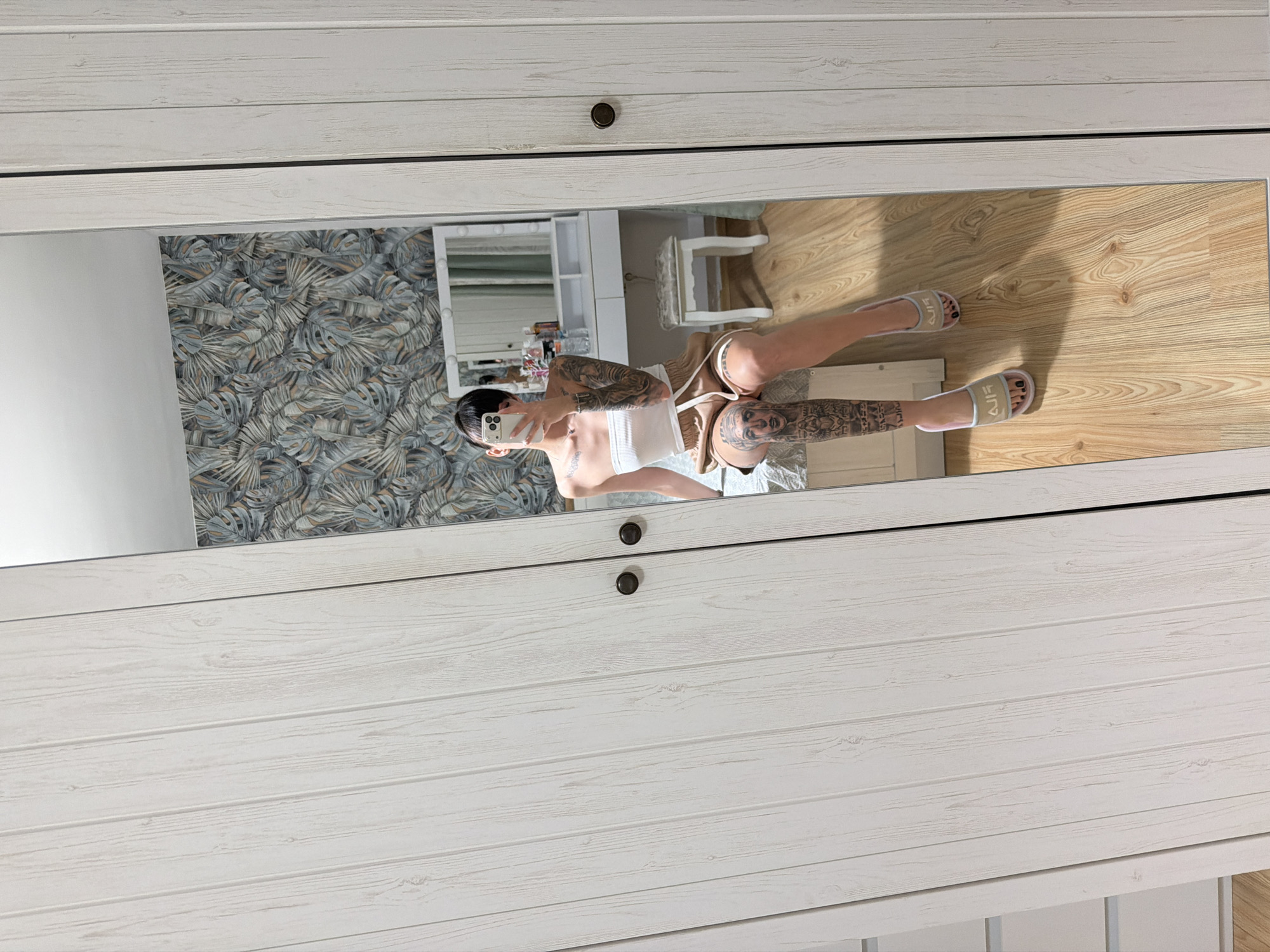 The image depicts a full-length mirror reflecting a woman sitting on a bed. The woman is wearing a white strapless top and beige shorts, and she has extensive tattoos on her arms and legs. She is holding a smartphone, presumably taking the picture. The room appears to be a bedroom, with a dresser and stool visible in the reflection. The walls are decorated with a tropical leaf pattern wallpaper. The floor is wooden. The mirror is positioned in front of a large, light-colored wardrobe with vertical paneling and dark circular handles. There are no identifiable landmarks or clues within the image that would indicate its location in Plovdiv, Bulgaria.