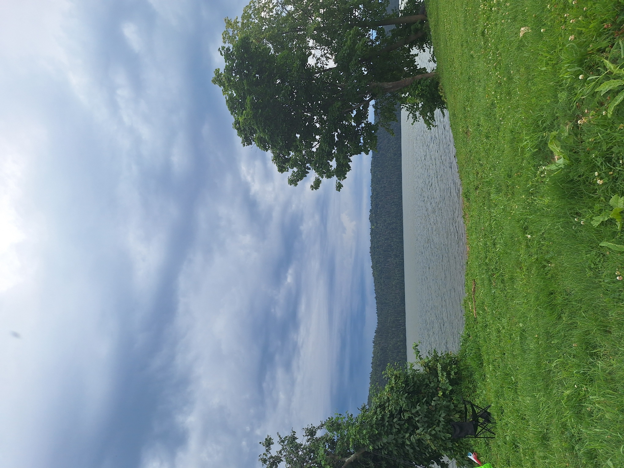 This is a landscape photograph of a lake under a cloudy sky. The foreground consists of a grassy area with a black folding chair and a small bush on the left. In the middle ground, a calm body of water stretches across the frame, with a tree-covered hillside forming the background. The trees on the right side of the frame are dense and green. The sky is filled with dark, dramatic clouds, suggesting overcast weather. The location is identified as Atamanovka, Russia.