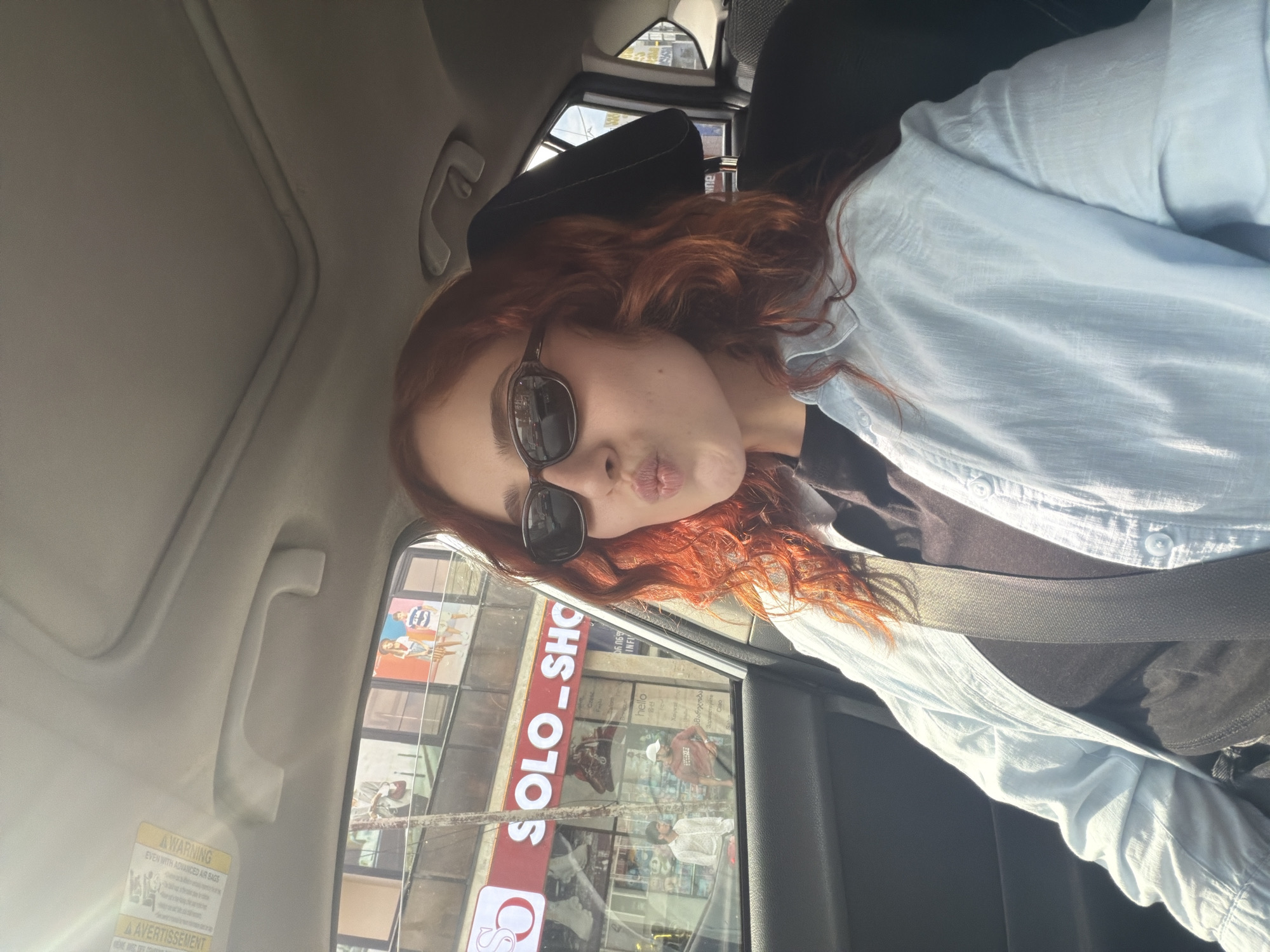 A portrait-style image, rotated 90 degrees counter-clockwise, features a person with long, wavy, reddish-auburn hair and dark sunglasses, making a playful "puckered lips" or "kiss face" expression. The person is seated in a car, likely in the passenger seat, wearing a light blue button-up shirt over a dark top, with a seatbelt visible across their chest.
The car's interior is light-colored, showing the ceiling, an overhead grab handle, and a sun visor with a "WARNING / EVEN WITH ADVANCED AIR BAGS / A AVERTISSEMENT" label. Bright daylight suggests a sunny day.
Through the car window, an urban street scene is visible, featuring buildings with large glass windows. A prominent red and white sign displays "SOLO_SHOP". Nearby, smaller text includes "690 YO", "INFI", "Cresc", and several greetings: "hello", "Χαιρετέ", "გამარჯობა" (Georgian), "Зарвест", and "Ciao". The presence of Georgian text strongly indicates the location is Tbilisi, Georgia. The overall scene captures a casual moment in a bustling city environment.