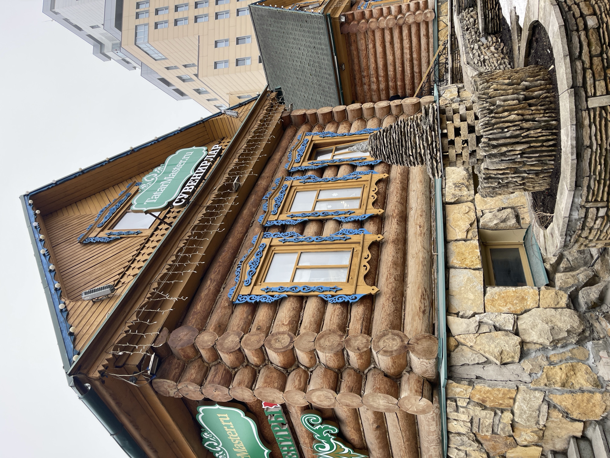 The image shows a building in Kazan, Russia, constructed of logs with carved and decorated windows and a sign that reads "TatarMaster.ru СУВЕНИРЛАР". The building also features stone construction on a portion of the facade, including an arched element. A modern, multi-story building is visible in the background. The sky is overcast.