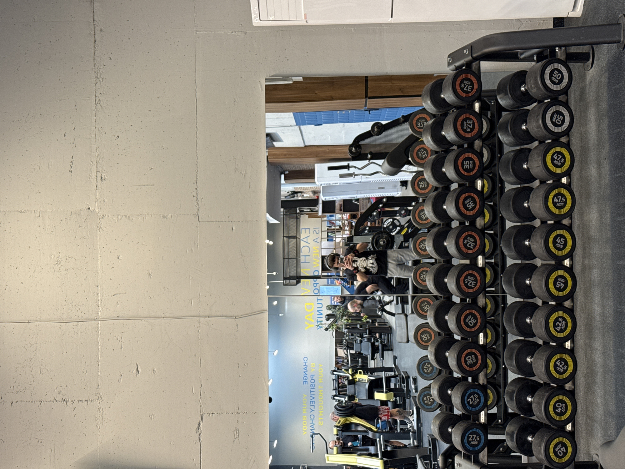 The image shows a gym's interior, reflected in a mirror. The gym contains various exercise machines and free weights, including dumbbells arranged on a rack. In the mirror's reflection, several individuals are exercising or using their phones. The gym equipment includes weight benches, cable machines, and other exercise apparatus. The background shows more of the gym's interior with visible fluorescent lighting. The mirror displays text, including "YOUR BODY, THOUGHT RUDY, CHANGE POSITIVITY, EACH IDEA". The location appears to be inside a gym in a city, likely Sofia, Bulgaria, though there's no explicit indication of the location within the image itself. The scene is well-lit and active, with people focused on their workouts.