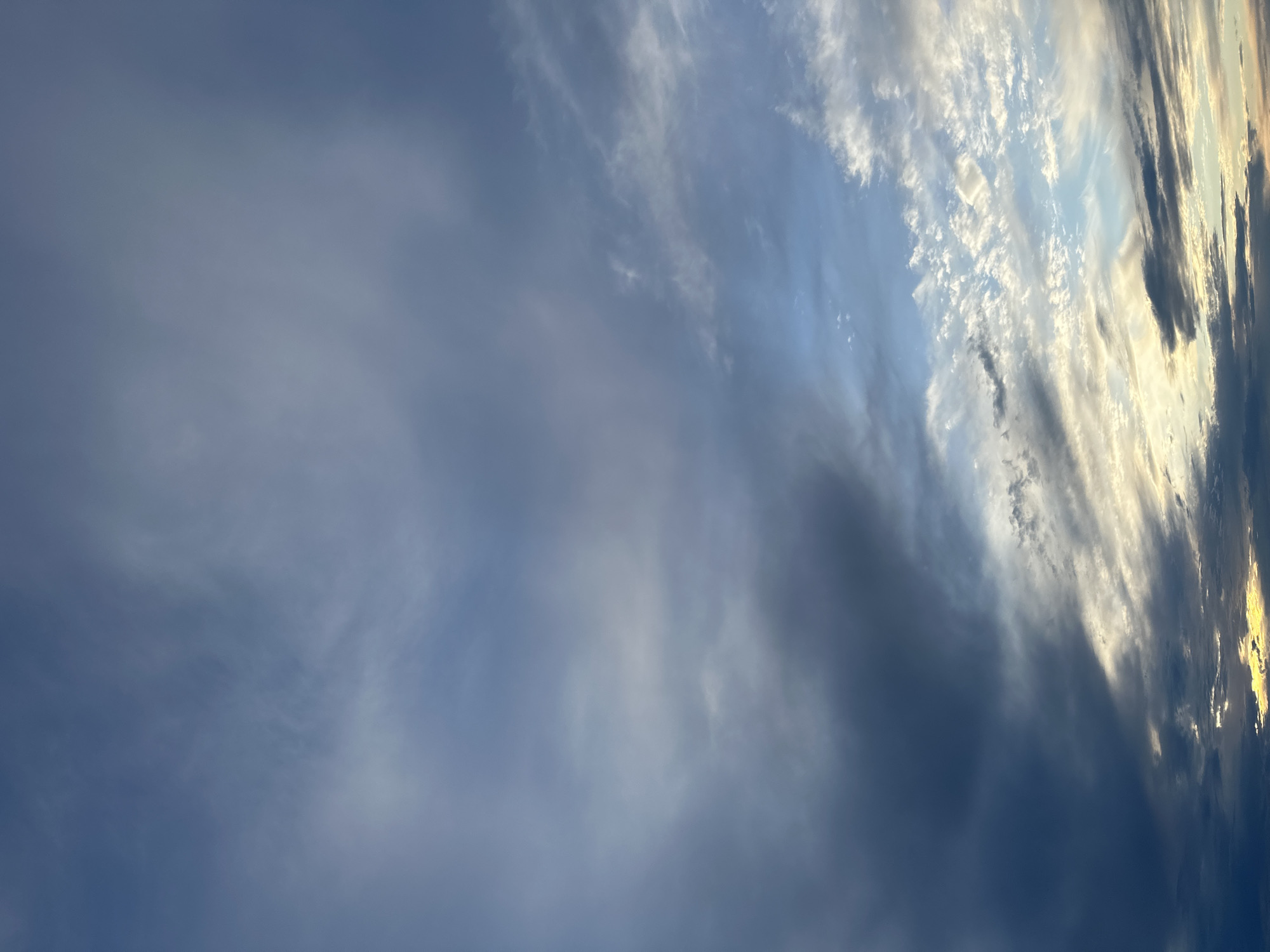 The image depicts a daytime sky in an unknown location. The sky is filled with a variety of clouds, ranging from thin, wispy cirrus clouds on the right side, to thicker, darker clouds in the middle and left side. The lighting suggests that the sun is low on the horizon, possibly during sunrise or sunset, casting a soft glow on the clouds. The overall impression is of a dynamic and changing sky, with different cloud formations and atmospheric conditions coexisting in the same scene.