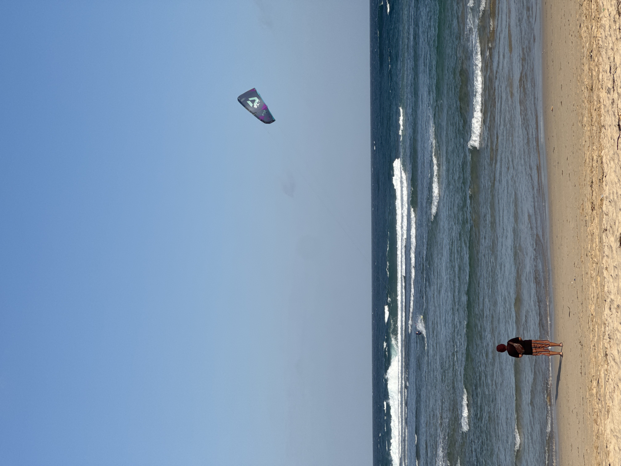 A wide shot of Kuta Selatan, Indonesia's coastline shows a clear blue sky above choppy ocean waves. In the foreground, a person stands on a sandy beach, facing the water. The individual wears a dark t-shirt, striped shorts, and a brown fanny pack. In the mid-ground, a kitesurfer is actively engaged in the sport. A large, multi-colored kite with the text "DUO" and "North" is visible in the upper right quadrant of the sky, indicating the kitesurfer's equipment. The ocean exhibits distinct lines of white-capped waves breaking as they approach the shore. The beach is composed of light-colored sand with some scattered debris. The overall scene suggests a bright, sunny day suitable for water sports.