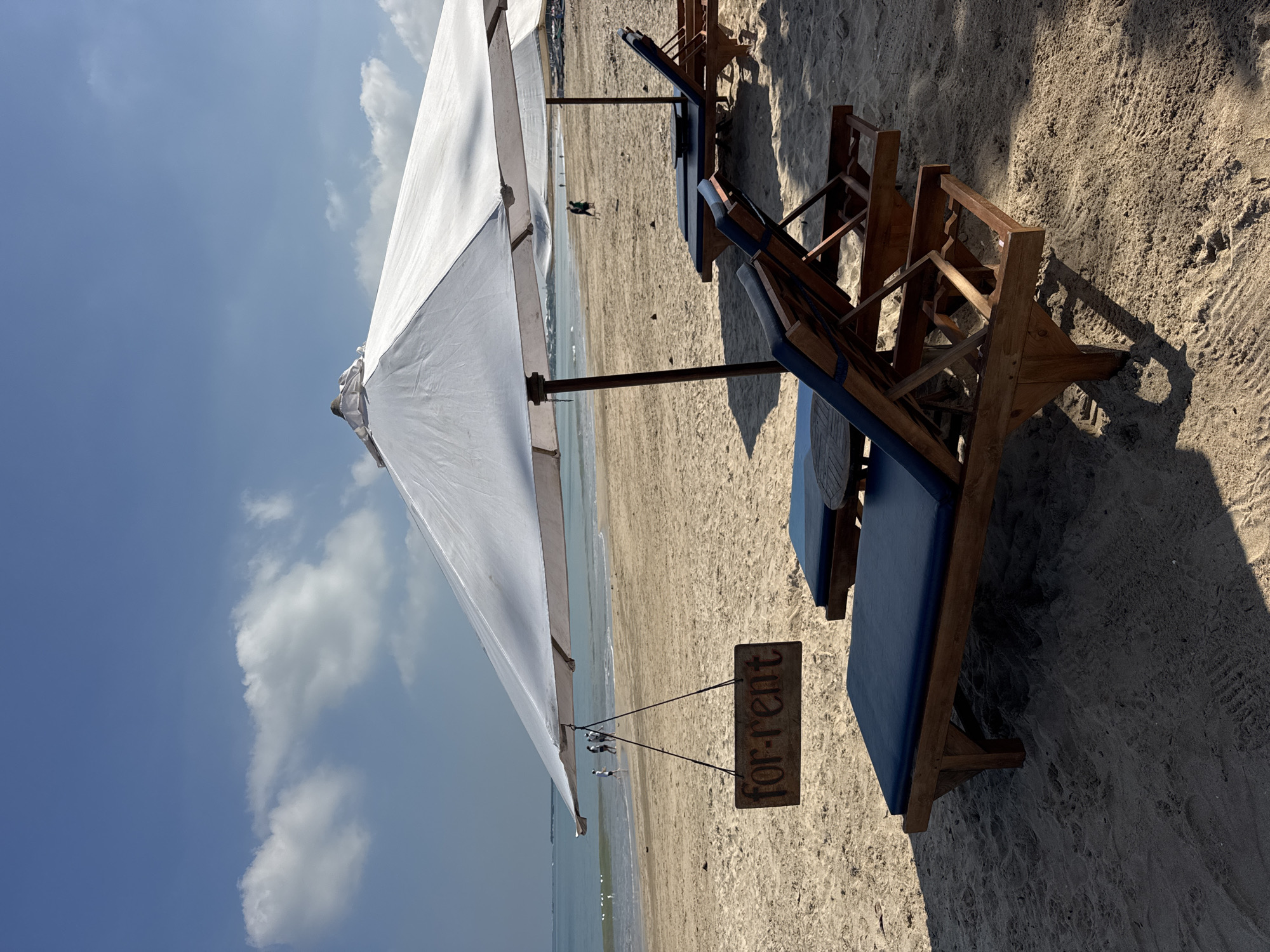 This image depicts a sunny beach scene in Kuta Selatan, Indonesia, looking towards the ocean. The foreground is dominated by a large white beach umbrella, under which several wooden beach chairs with blue cushions are arranged on the sand. A rustic wooden sign, clearly reading "for-rent," hangs from the pole of the prominent umbrella, indicating the availability of the equipment.
Bright sunlight illuminates the scene, casting distinct, long shadows of the umbrellas and chairs onto the textured sand, suggesting either morning or late afternoon. Beyond the immediate beach furniture, the sandy shore stretches towards the calm, light blue water of the ocean. Several small figures are visible in the distance near the waterline, presumably beachgoers enjoying the tranquil environment.
The sky is a clear, vibrant blue with scattered white clouds, reinforcing the sunny and pleasant weather conditions. The overall setting portrays a typical tropical beach destination, inviting relaxation and leisure.