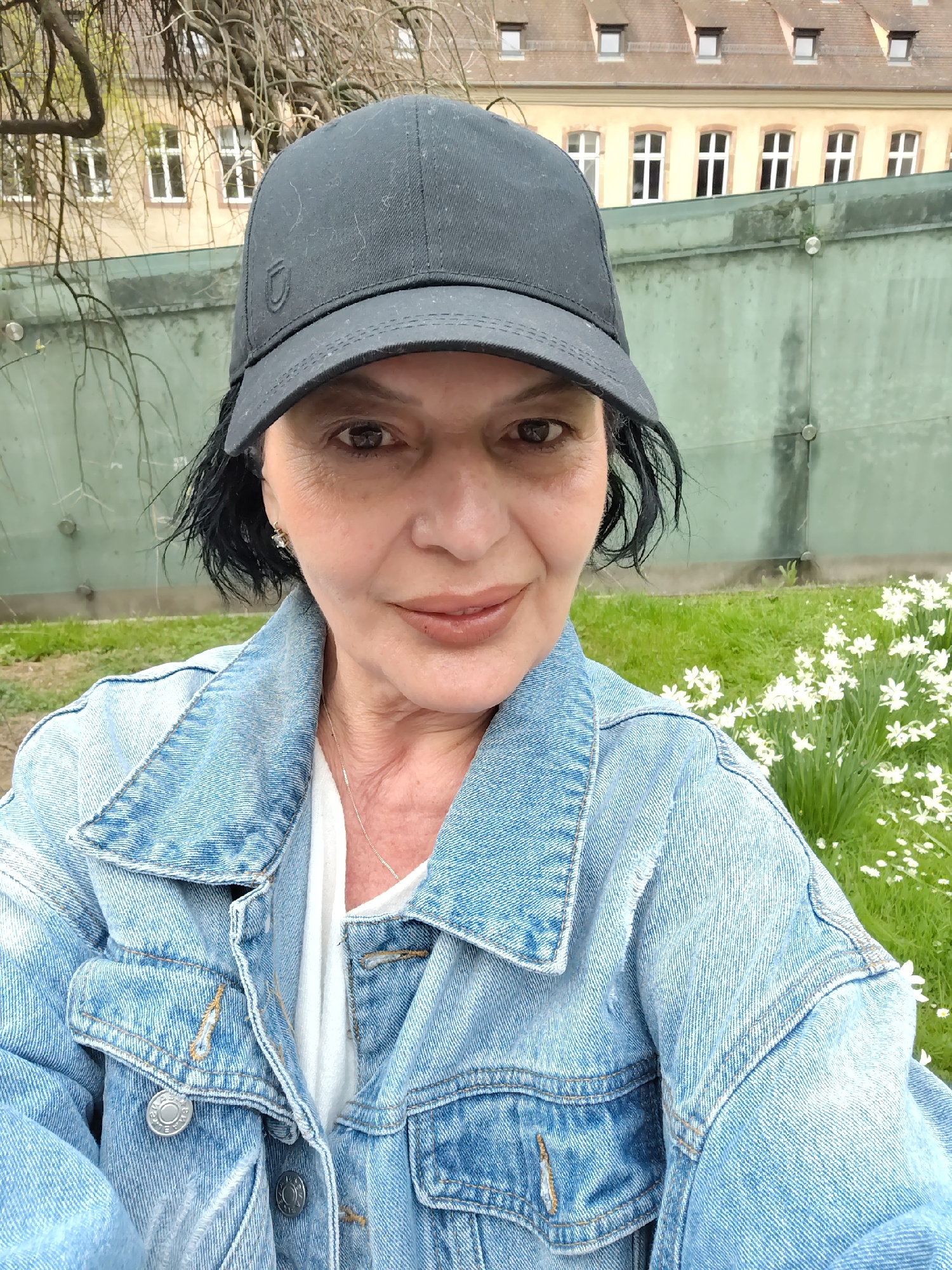 A middle-aged woman wearing a denim jacket and a black baseball cap takes a selfie. She is positioned in front of a green wall, with a grassy area featuring white flowers to her right. In the background, a building with multiple windows and dormers is visible. The location is Strasbourg, France.