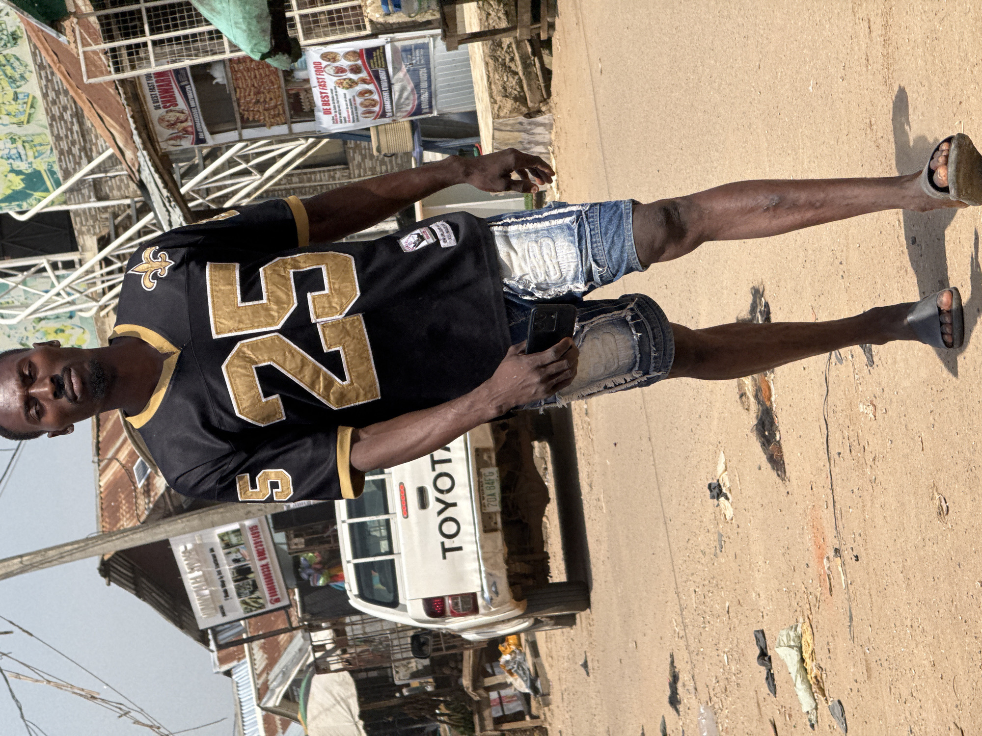 A full-shot, outdoor, eye-level, slightly high-angle photograph captures a Black male, appearing to be in his 20s or 30s, walking towards the camera on a dirt road. He is wearing a black and gold New Orleans Saints football jersey with the number 25 prominently displayed in gold, and distressed denim shorts. He is holding a black smartphone in his right hand and wearing dark grey slide sandals. His facial expression is neutral, and he is looking directly at the camera.
The background depicts a bustling street scene in Abuja, Nigeria. Numerous shops with corrugated metal roofs line the street, some with signs advertising food and services, such as "DE BEST FAST FOOD SHAWARMA" and "DE BEST FAST FOOD." A white Toyota pickup truck is parked on the left side of the road, with its license plate visible. The ground is a dusty, dirt surface with scattered debris, including plastic bags and fabric scraps. The overall environment suggests a typical street scene in a commercial area of Abuja. The lighting indicates a bright, sunny day.