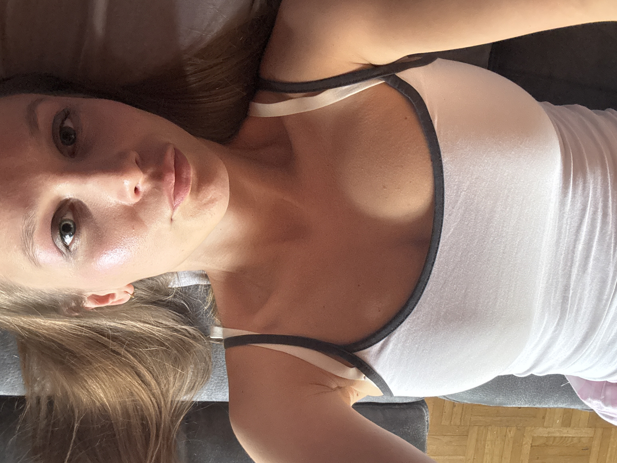 The image shows a close-up, overhead view of a young woman lying on a gray couch. She has long, blonde hair spread out around her head. She is wearing a white tank top with dark gray trim. The woman is looking directly at the camera with her eyes open. Her face is illuminated by natural light, highlighting her skin. The background consists of the gray couch and a portion of a wooden floor with a parquet pattern. There are no identifiable landmarks or contextual clues that would suggest a specific location within the city of Überherrn, Germany. The scene is static and there is no discernible action taking place.
