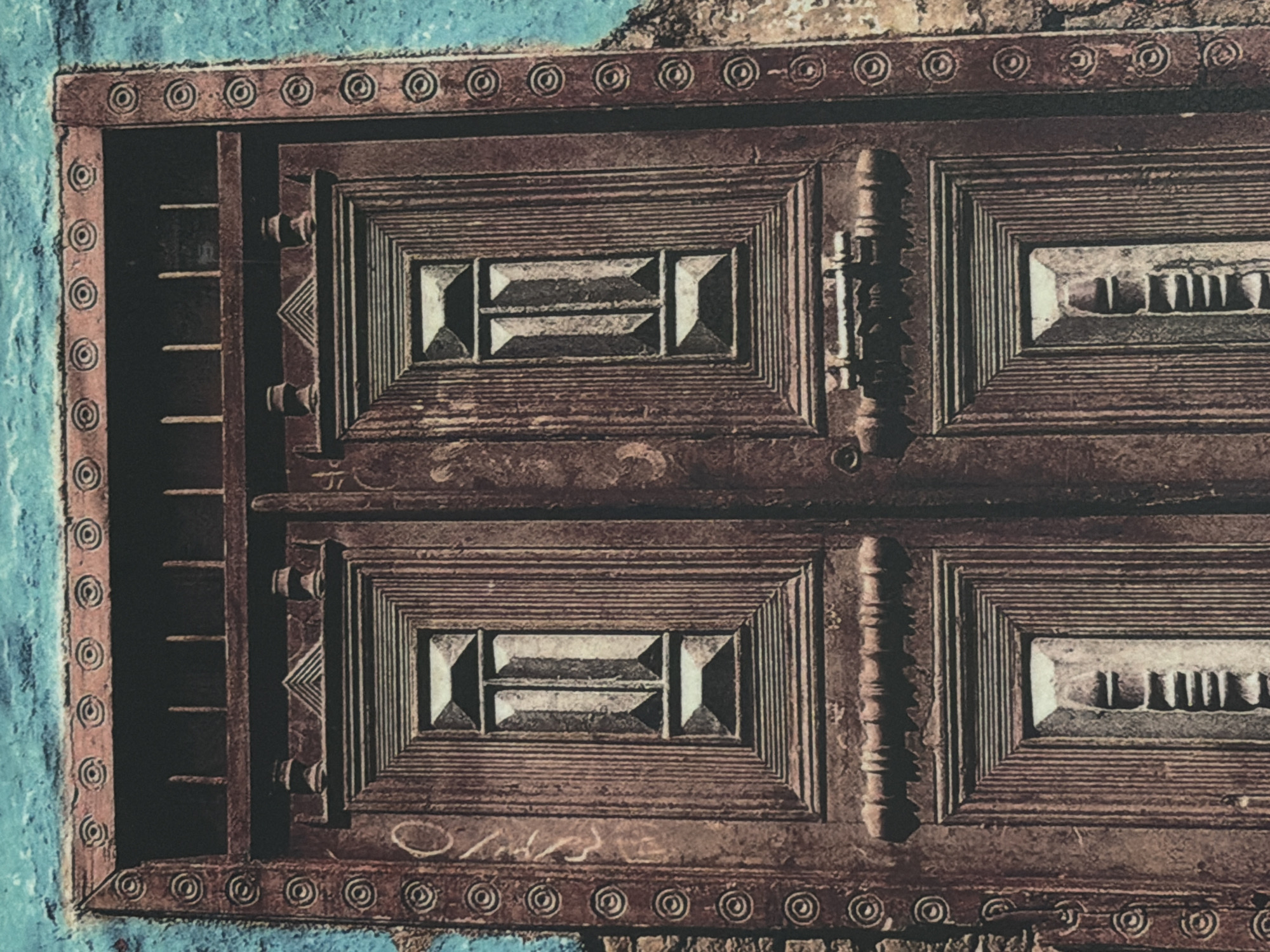 The image shows a close-up, front-facing view of a large, weathered wooden double door with intricate carvings. The door is a dark reddish-brown and appears to be made of aged wood, showing signs of wear and possibly some cracking. It is set within a textured, turquoise-colored wall on the left and a stone wall on the right.
The door is divided vertically into two panels, each featuring a central rectangular inset with a decorative, multi-layered geometric design. These insets are framed by a series of concentric rectangles and vertical lines. At the top of each door panel, there are two small, decorative finials. Above the doors, there is a horizontal beam with vertical slats, suggesting an upper section or a decorative element. The top edge of the door frame is adorned with a repeating pattern of circular motifs.
The left side of the door has some faint, light-colored graffiti. The overall impression is one of age, history, and ornate craftsmanship. The image does not depict any people or any immediate action. The environment is architectural, with the focus solely on the door and its immediate surroundings. The location of this door in Kyiv, Ukraine cannot be determined from the provided image.