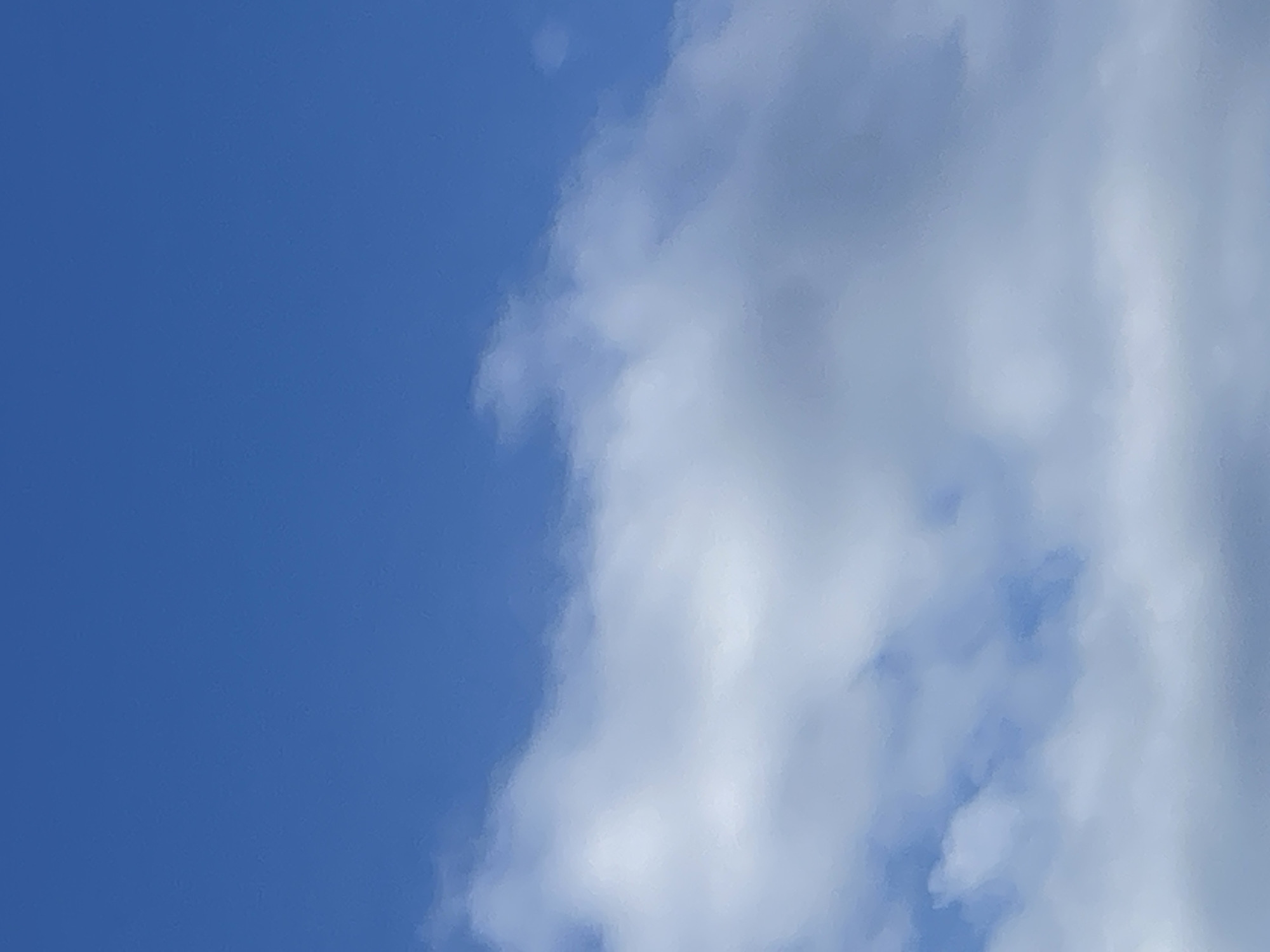The image depicts a bright blue sky with scattered, soft white clouds. The clouds are primarily located in the lower half of the frame, appearing somewhat wispy and diffused, suggesting either a sunny day with light cloud cover or possibly a hazy atmosphere. The upper portion of the image is dominated by uninterrupted blue sky. There are no visible structures, people, or identifiable landmarks within the frame. The lighting suggests it is daytime. The context of Sviatopetrivske, Ukraine, implies this is an outdoor scene, likely a view looking upwards.