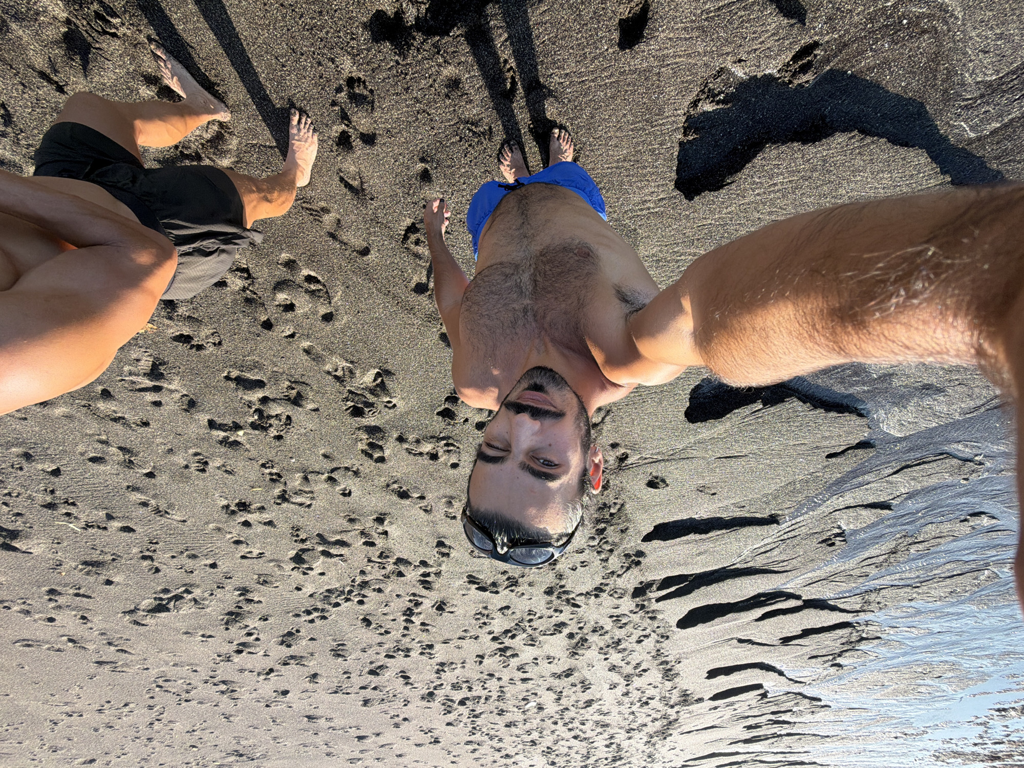 A high-angle, wide shot captures a man taking a selfie on a beach in Mengwi, Indonesia. The man, with dark hair and a mustache, winks at the camera, wearing blue swim trunks and sunglasses pushed up on his head. His bare chest is visible. Another person's arm and shoulder are visible in the lower right corner. The sand is dark and wet, with footprints scattered across it, suggesting it's a beach near the water's edge. The water is visible on the left side of the image, with ripples and patterns formed on the wet sand. The lighting indicates it's daytime, possibly late afternoon due to the long shadows cast by the figures.