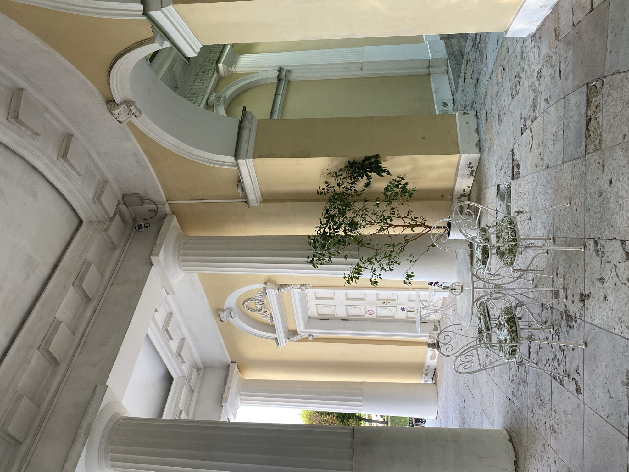 This image captures an outdoor courtyard with classical architectural elements, likely in Saint Petersburg, Russia. The scene features a colonnade with large, fluted white columns supporting a pale yellow ceiling with ornate decorative molding. Arches and decorative elements in white plaster add to the neoclassical aesthetic.
The ground is paved with large, irregular stone tiles, some of which appear worn and chipped. In the foreground, there is a white metal bistro set consisting of a table and two chairs. The chairs have intricate scrolled metalwork and cushions with a green and white floral pattern. A potted plant with green leaves is placed near the table.
In the background, a white double door with decorative paneling is visible. Above the door, there are more architectural ornaments. A security camera is mounted on one of the walls. The lighting suggests it is daytime, with the sun casting shadows and illuminating the scene. The overall atmosphere is serene and somewhat understated, hinting at a quiet moment in a historic setting. There are no people visible in the image, but the presence of the bistro set suggests it's a place for relaxation or enjoying a drink outdoors.