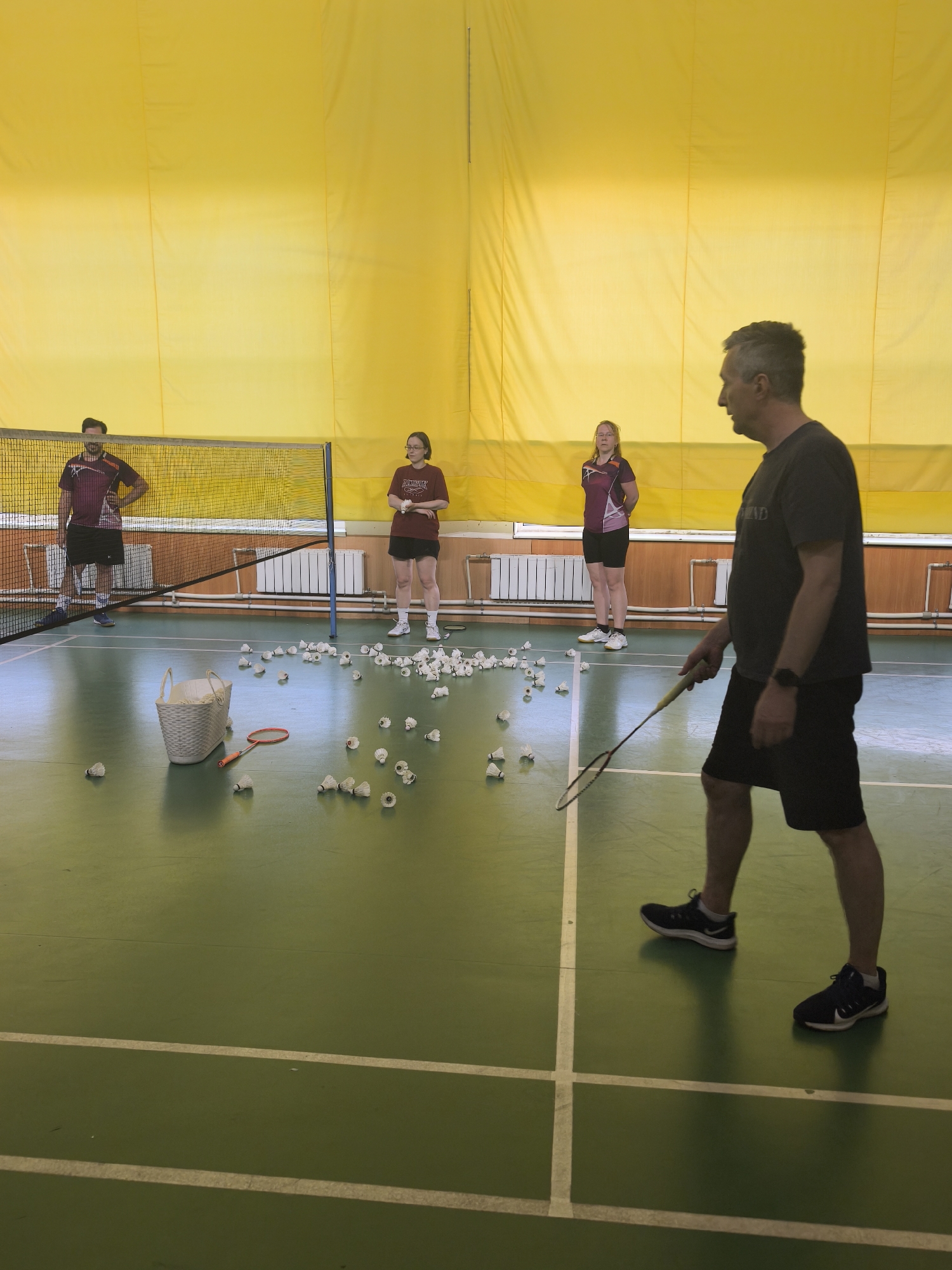 The image displays an indoor badminton court, likely within a school gymnasium or a community sports club in Saint Petersburg, Russia. The exact address or specific facility name cannot be determined solely from the visual information provided. The environment features bright yellow, vertically seamed padded walls extending from mid-height to the ceiling. Below the yellow padding, a light brown wooden or laminate wainscoting runs along the lower wall, with white finned radiators affixed to it. The floor is a distinct olive-green synthetic sports surface with clear white boundary lines delineating a badminton court. The lighting appears bright and even, suggesting overhead artificial illumination. Four individuals are present on and around the court, engaged in what appears to be a badminton training session or casual play. In the foreground, on the right side of the frame, a middle-aged man with short, graying hair and stubble is walking towards the left. He wears a dark (black or dark gray) short-sleeved t-shirt with a light logo (partially visible as "BLEND"), dark (black) athletic shorts, white socks, and black athletic shoes with white soles and a white swoosh logo. He has a dark wristband or watch on his left wrist. He holds a badminton racket in his right hand, with the strings facing downwards. To the left of the net, a younger man with short dark hair and a beard stands observing. He wears a maroon or dark purple short-sleeved athletic shirt with light design elements, black athletic shorts, blue socks, and light blue and gray athletic shoes. His right hand is on his hip, and his left arm is relaxed by his side. Behind the net, towards the right, a woman wearing glasses stands. She has light brown hair pulled back and is dressed in a dark red or maroon short-sleeved t-shirt with a light logo on the chest, black shorts, white socks, and white athletic shoes with dark soles. She stands with her hands clasped in front of her, observing the court. Further in the background, to the right of the woman in glasses, another woman stands near the yellow wall. She has light brown hair, wears a purple or magenta short-sleeved athletic shirt with light design elements, black shorts, white socks, and white athletic shoes. Her hands are clasped behind her back as she observes the scene. On the court floor, primarily on the side closest to the camera and near