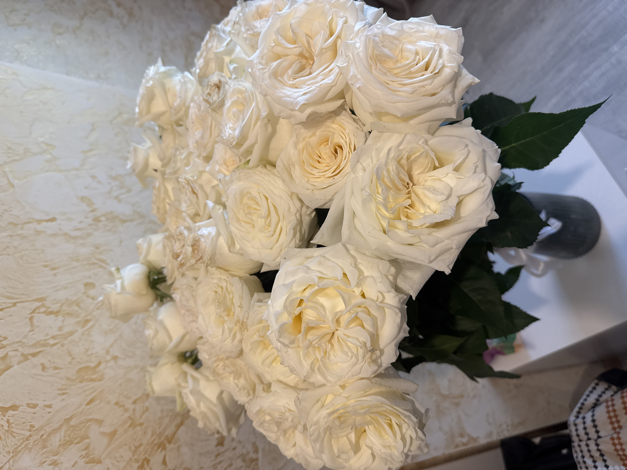 The image depicts a bouquet of white roses, arranged in a dense cluster. The roses are mostly in full bloom, revealing their petal structure. Green leaves can be seen interspersed among the roses. The bouquet sits on a surface, with patterned fabric visible in the background. A vase, partially obscured, is present. The location is Unknown, Unknown. No people are visible. The scene focuses on the floral arrangement.