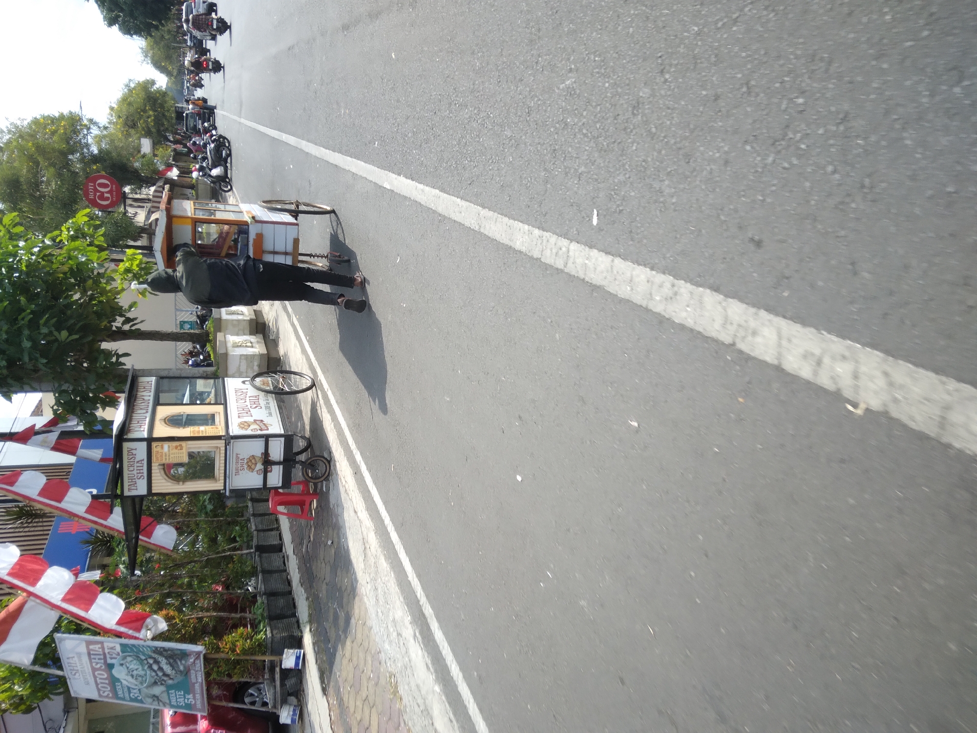 In Purwokerto, Indonesia, a person is pushing a food cart with the label "TAHU CRISPY SHIA" on the side, down a road. The cart is a mobile vendor, equipped with a canopy and a glass display case. The individual is wearing dark clothing and appears to be working. In the background, there are other vendors, parked motorcycles, cars, and trees lining the street. The scene suggests a busy street environment, likely in a commercial or populated area. The presence of Indonesian flags also hints at a nationalistic or celebratory context.