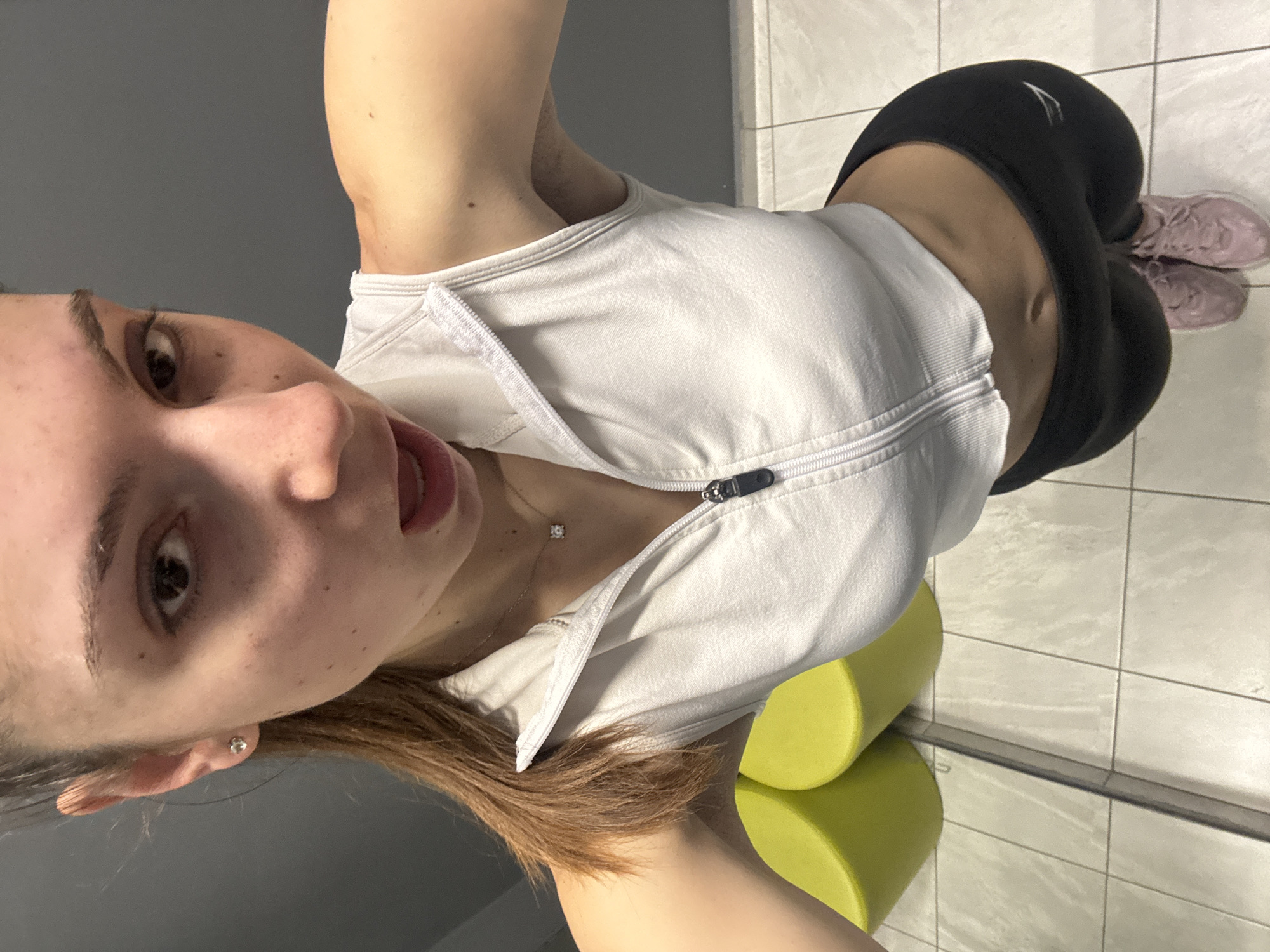 An indoor full-body selfie shows a young woman rotated 90 degrees clockwise, striking a playful pose, likely in a gym or fitness center in Varna, Bulgaria. She has light brown hair, an oval face, and is looking directly at the camera with her mouth slightly open and tongue partially extended. She wears a white sleeveless athletic top with a partially unzipped front, revealing a delicate silver necklace with a small, sparkling pendant. A small silver stud earring is visible in her left ear. She is dressed in form-fitting black athletic leggings, on which a white logo resembling the 'Gymshark' brand is visible near her left hip. Her feet, visible at the bottom of the frame, are clad in light pink athletic shoes. The background features a plain grey wall and a light-colored tiled floor. Two bright lime green cylindrical objects, possibly foam rollers, are situated on the floor, part of which is reflected in a mirror running along what would be the bottom of a correctly oriented image. The lighting is bright and even, suggesting a well-lit indoor environment. The overall scene captures a casual moment, perhaps during or after a workout.