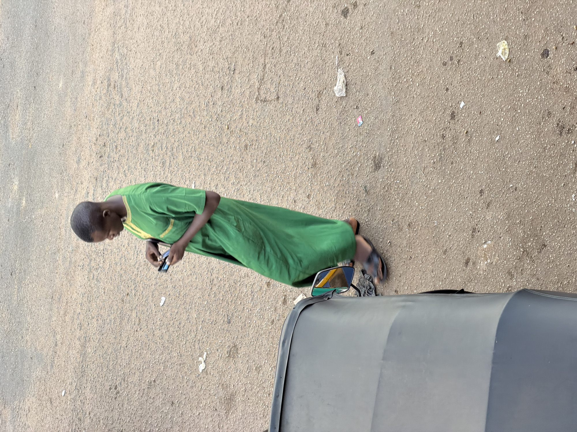 A young boy is standing on a dirt road in Bukuru, Nigeria, looking at his mobile phone. He is wearing a green robe with yellow trim. A dark-colored vehicle, possibly a three-wheeled auto-rickshaw, is parked next to him, with its side mirror visible. The road is covered with small stones and pebbles, and there is some scattered litter. The boy appears to be alone, and the overall impression is one of everyday life in a busy, dusty environment.