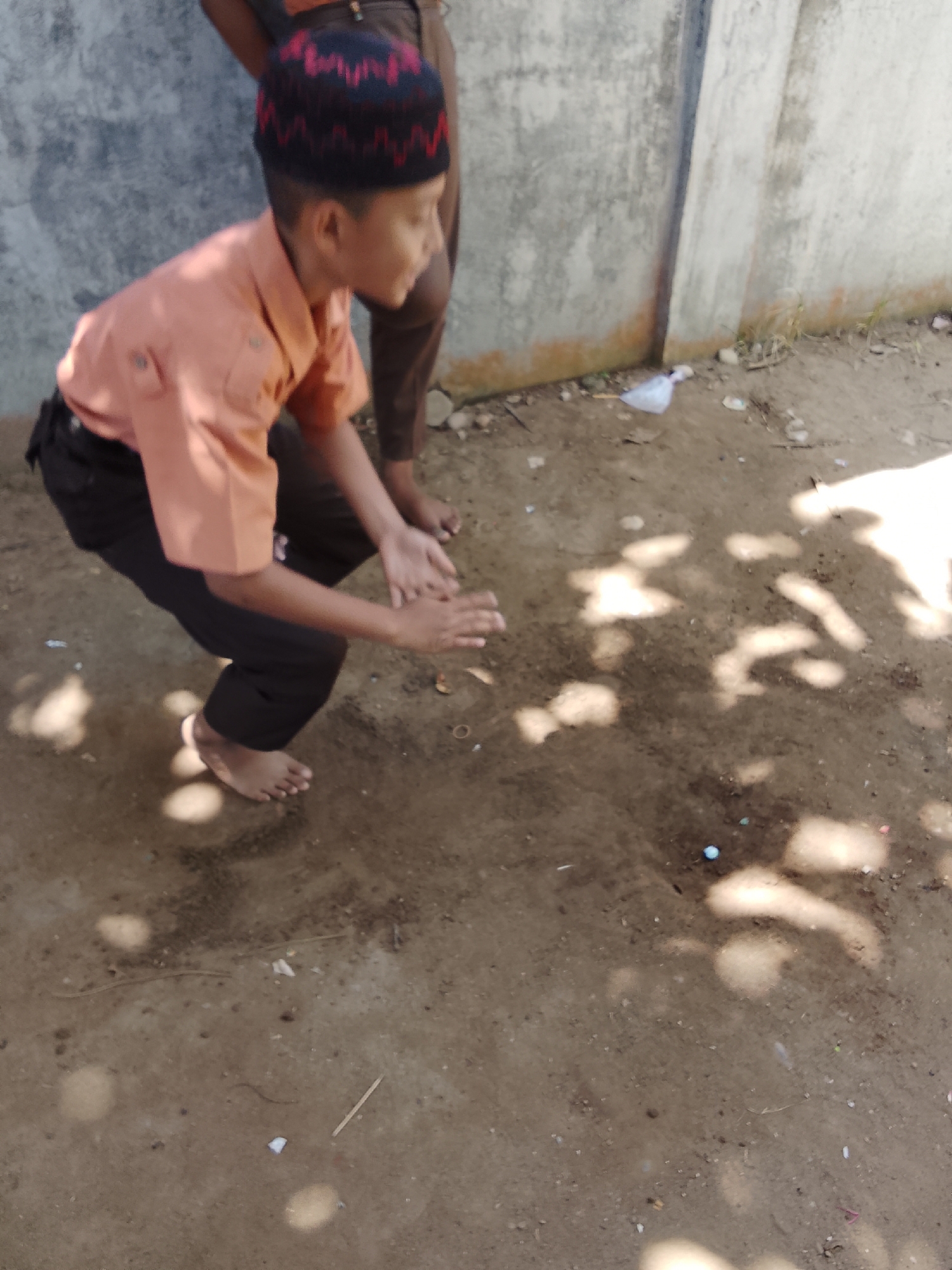 A young boy wearing a peach-colored shirt, black pants, and a black and red patterned cap is crouched on the ground. His hands are in front of him, palms up, as if he is about to catch something or is interacting with something on the ground. The ground is dirt and appears to be in an outdoor area, possibly a yard or street. There are shadows cast on the ground from what appears to be overhead foliage. A wall with a rough texture is visible in the background. The location is identified as Baratpayaitek, Indonesia.