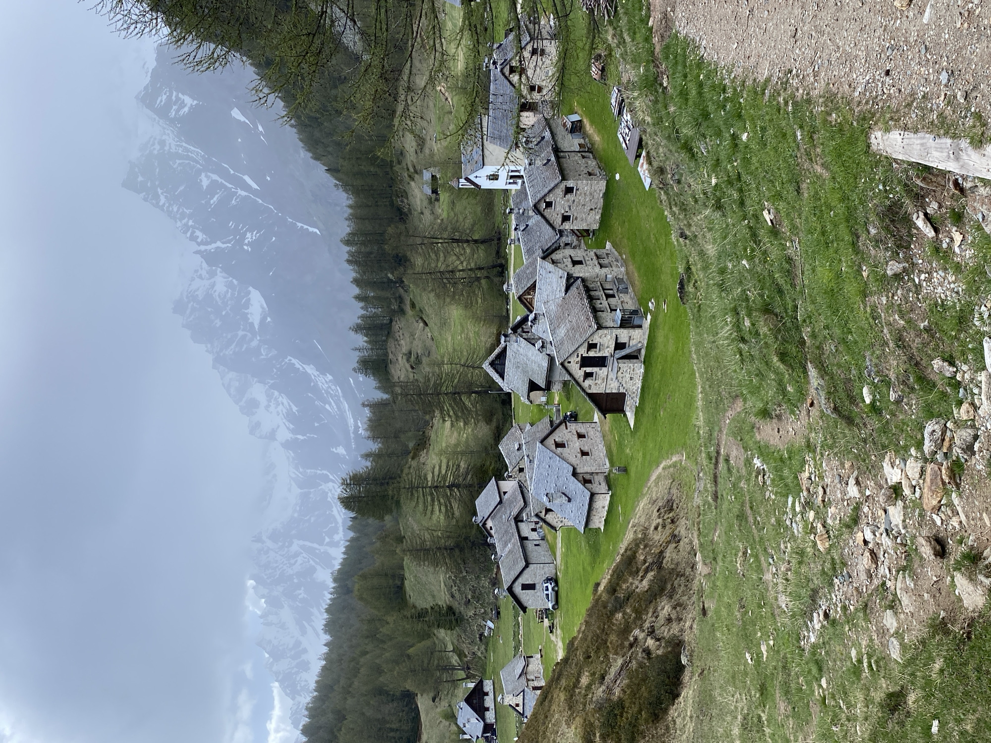 The image depicts an alpine village composed of stone buildings with slate roofs nestled in a green valley. The structures appear to be traditional in style, exhibiting a rustic charm. The location is identified as Croveo, a frazione (hamlet) within the comune (municipality) of Baceno, Verbano-Cusio-Ossola, Piedmont region of Italy.
The mountains, partially obscured by clouds, serve as a dramatic backdrop to the village. Trees line the valley side. There are no people visible in the scene. The overall impression is one of tranquility and seclusion in a mountainous environment.