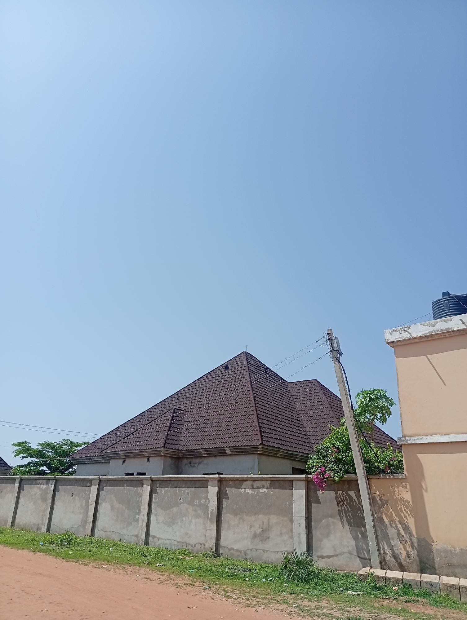 The image depicts an outdoor scene under a clear blue sky, possibly in Wauro Jabbe, Nigeria. In the mid-ground, a large building with a prominent brown, tiled, multi-peaked roof is visible, bordered by a textured concrete wall. The wall is partially covered by green vegetation. A concrete utility pole with electrical wires runs diagonally across the right side of the frame. To the right of the pole, a portion of a tan-colored building with a concrete lip and a black water tank on top is visible. The foreground shows a dirt road with scattered green grass along its edge. The overall impression is one of a residential or commercial area under construction or recently completed, given the unfinished appearance of some structures and the presence of utility infrastructure.