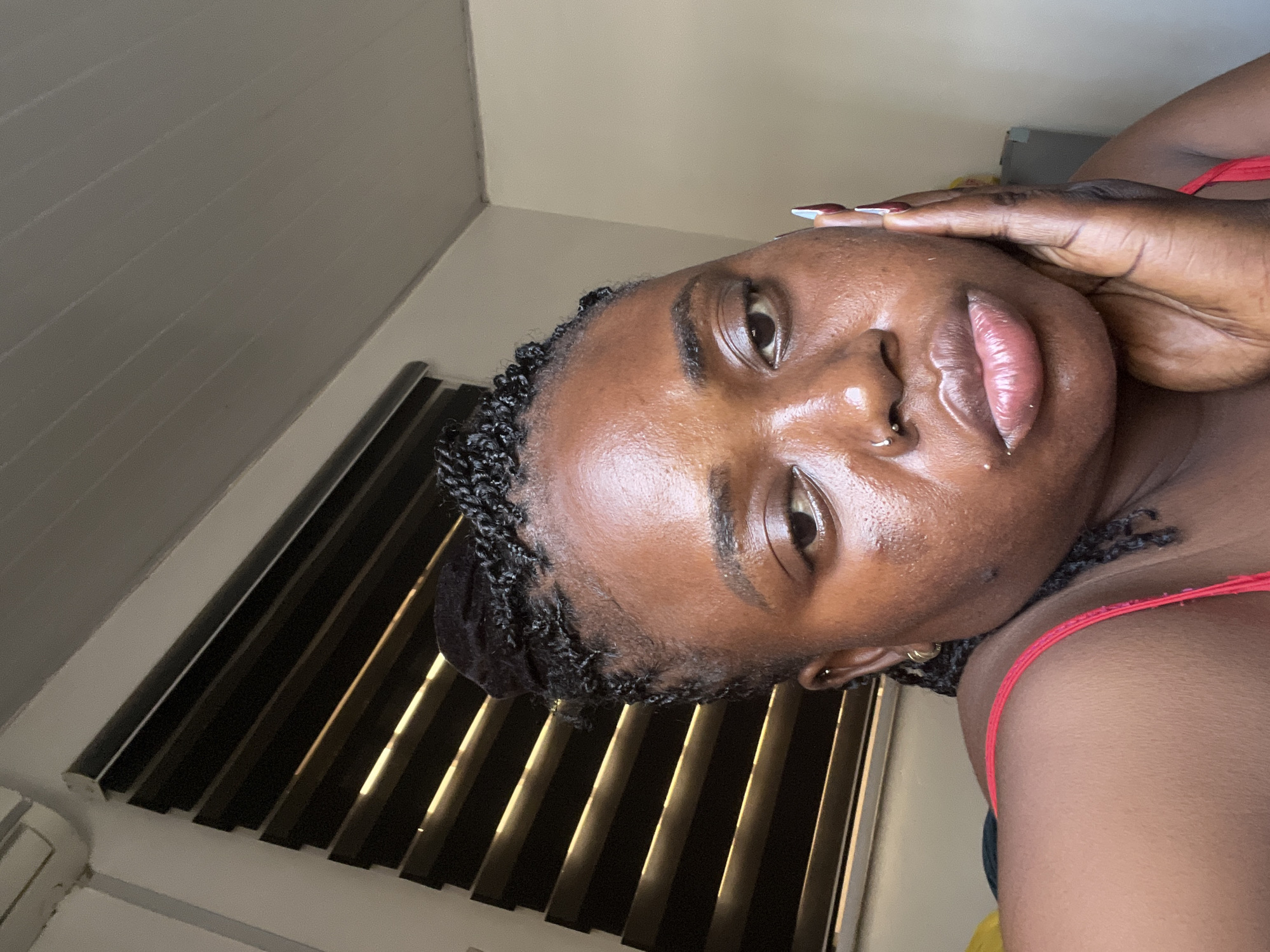 A close-up, waist-up portrait shows a young Black woman with her face turned slightly towards the camera, resting her right hand on her cheek. She has dark skin, dark braided hair pulled up, and a nose ring. She is wearing a red tank top. The background features a window with horizontal blinds, an air conditioner unit, and a plain wall. The lighting suggests the photo was taken indoors during the day. This image does not contain any information that would allow for its location to be identified as Abuja, Nigeria.