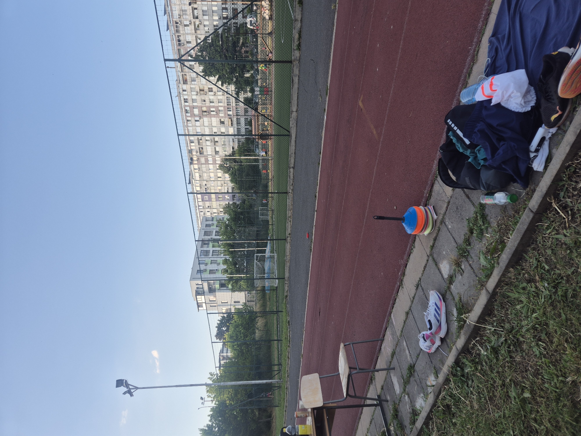 An outdoor athletic track and field scene is depicted under a clear blue sky. In the foreground, a red track runs diagonally across the image, bordered by a concrete walkway and grass. On the walkway, a pair of white athletic shoes with purple and blue accents are placed. Further to the right, a stack of colorful training cones is visible. Adjacent to the cones, a pile of sports equipment lies on a blue mat, including a black backpack, a dark blue shirt or towel, a white plastic bottle, and other garments. A single black athletic shoe with an orange sole is also present. To the left, a table holds a yellow bottle and a can. A simple wooden chair with a metal frame sits beside the table. In the middle ground, a chain-link fence separates the track from a grassy area and a sports field. Beyond the fence, there are buildings, trees, and people participating in sports. In the background, large apartment buildings with numerous windows and air conditioning units are visible. A tall lamppost with a light fixture stands on the left side of the frame.