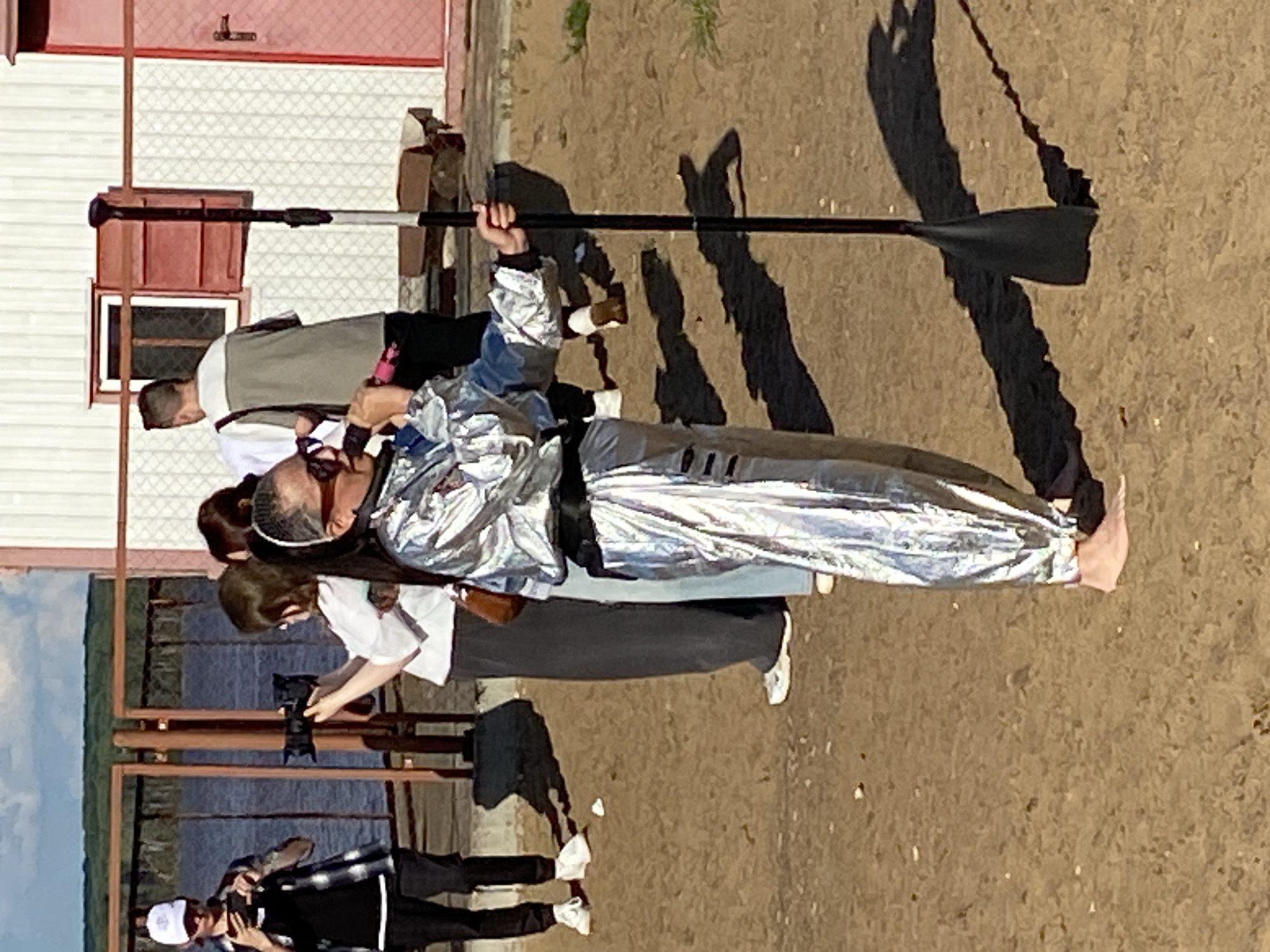In Chaikovsky, Russia, on a sandy, outdoor, waterfront area, a person wearing a shiny silver outfit with a black belt and dark pants is holding a microphone and speaking. The person has sunglasses on and dark hair pulled back. They are barefoot. To the right of the speaker, a tall, black pole with a paddle at the bottom is standing upright. In the background, a white building with red trim and windows is visible, as well as a fence and a body of water. Several other individuals are present in the scene, some holding cameras and appearing to film. The sun is casting shadows on the sand.