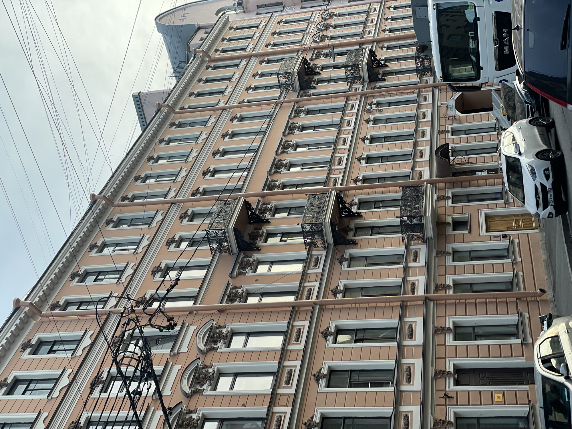 This image captures a multi-story residential or commercial building with a light peach-colored facade and white architectural trim, characteristic of Saint Petersburg, Russia, confirmed by the "178" region code on a visible license plate. The building features tall, narrow windows, many with decorative surrounds, and multiple floors equipped with ornate black wrought-iron balconies. Vertical drainpipes, matching the building's color, descend along the facade. Numerous overhead power lines crisscross the upper portion of the image against an overcast, light blue-grey sky.
At street level, several vehicles are parked or in motion. A white Hyundai Tucson SUV with license plate C 394 KB 178 is prominently visible. Partially visible vehicles include a dark sedan, a silver sedan, a red/dark car in the foreground, and the front portion of a white MAN truck on the right. A dark brown wooden door and a lighter wooden door are visible at ground level. An entrance with a sign reading "HELIX" is also present further down the street. The overall scene depicts an urban streetscape with no discernible people.
