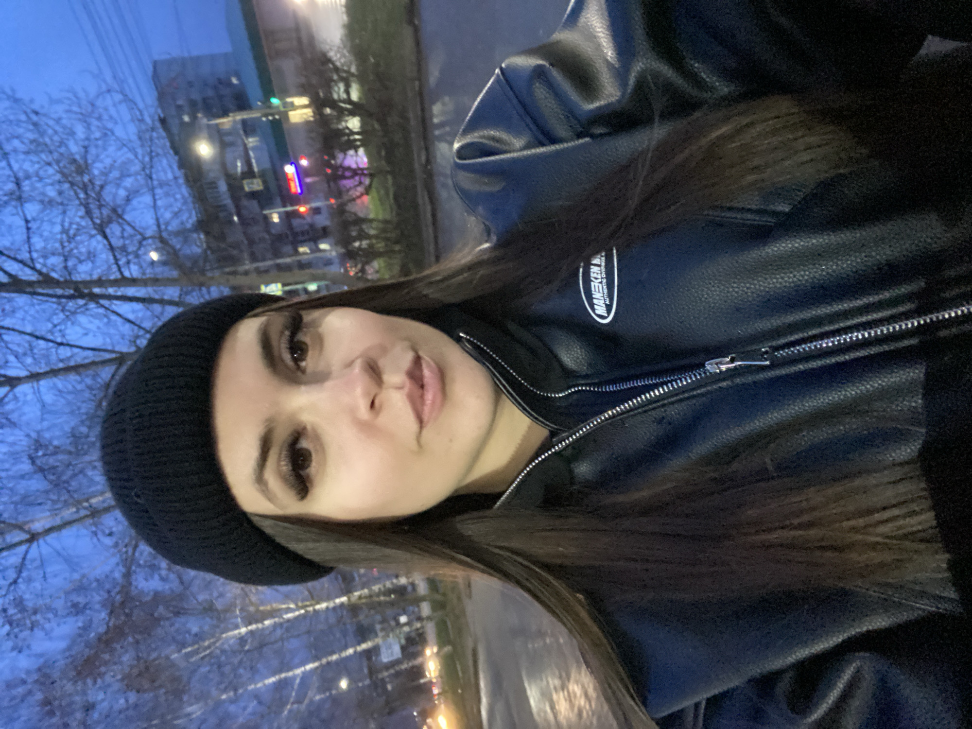 A woman with long brown hair, wearing a black beanie and a black leather jacket with a visible logo reading "MANEKEN B," is taking a selfie. The background shows a cityscape at dusk or early evening. Bare trees are visible to the left, and in the distance, buildings with lights suggest a location in Chaikovsky, Russia, possibly near a street. The overall impression is of an urban environment in a colder climate.