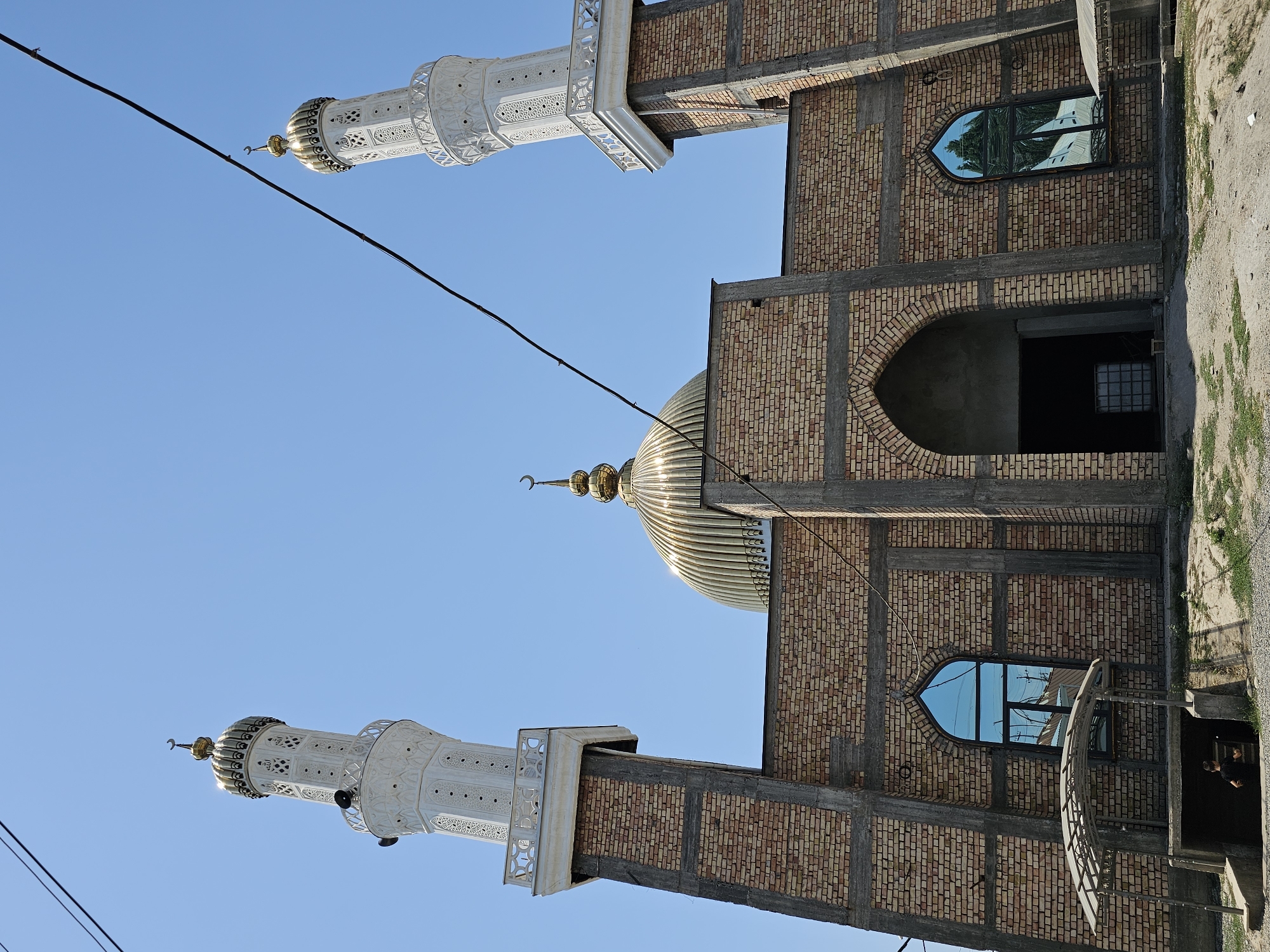 This is a low-angle, full shot of a mosque under construction in the city of Özgön, Kyrgyzstan. The building is made of light-colored brick with concrete structural elements visible. Two tall, white minarets flank a central golden dome. The minarets have decorative patterns and balconies, topped with crescent moons. The dome is ribbed and gleams in the sunlight, also topped with a crescent moon. An arched entrance to the building is visible, as well as two tall, arched windows with reflective blue glass. Power lines cross the sky above the mosque. The ground in front of the mosque is unpaved and scattered with gravel and some vegetation. A small, covered shelter is located to the left of the building, and a person wearing a dark shirt is visible descending into a dark opening on the left side of the frame.
