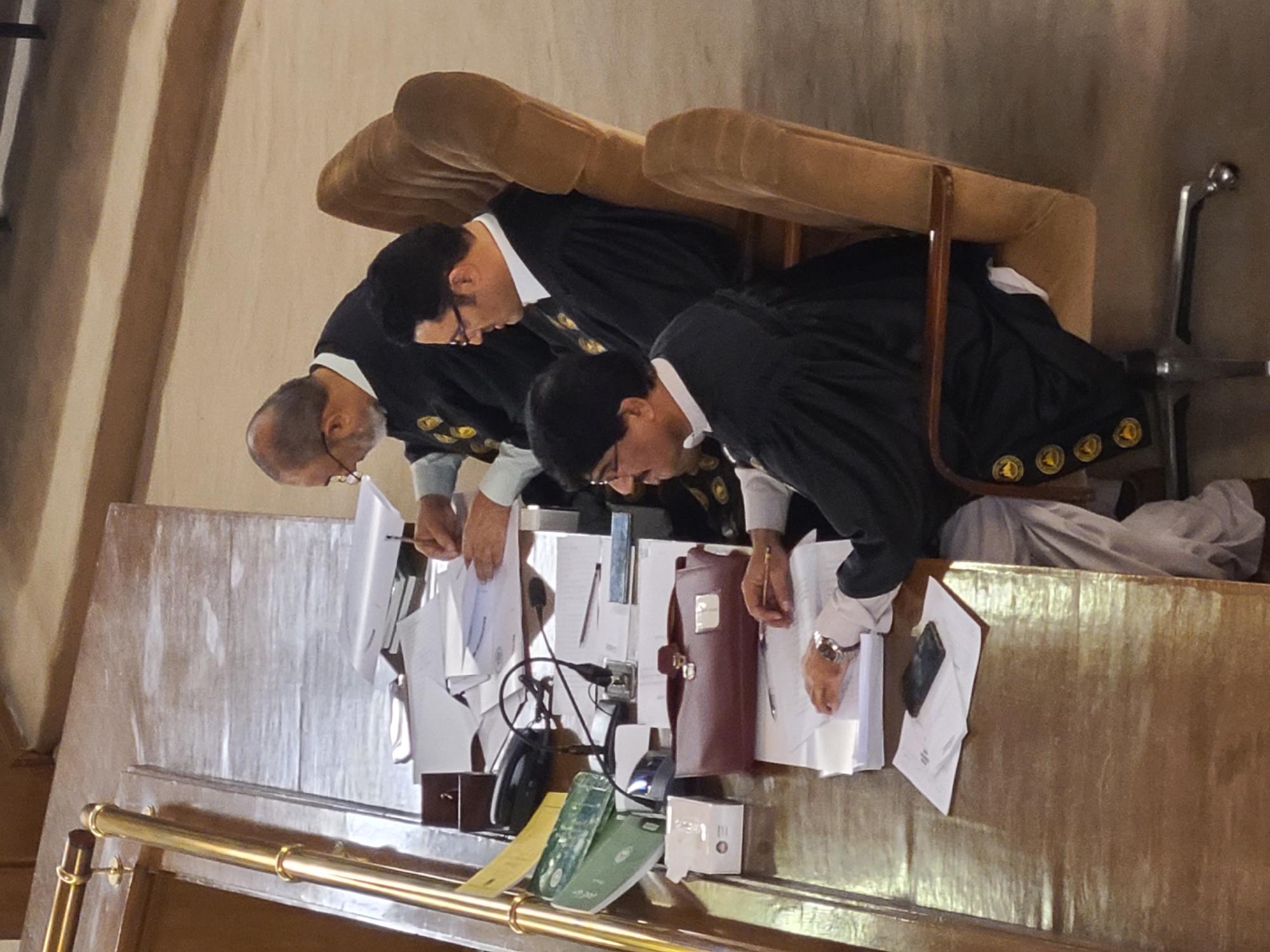 Three men in black robes with gold trim and white shirts are seated at a long wooden table covered in papers, a briefcase, electronics, and other items. The men are bent over the table, seemingly writing or reviewing documents. They are seated in chairs with brown upholstery. The location is likely a courtroom or legal setting in Quetta City Tehsil, Pakistan.