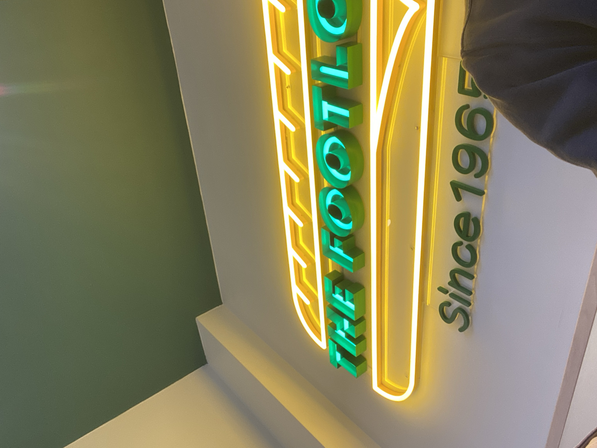 A vertically-oriented shot reveals a green and yellow neon sign, partially obscured by a person's shoulder and arm in the upper right. The sign reads "The Foot Look Since 1965". The sign is affixed to a white wall, adjacent to a green wall in the background. A portion of the wall beneath the sign appears cream-colored. There is a bright spot of light near the top of the image, on the green wall. The context suggests the location of Varna, Bulgaria.
