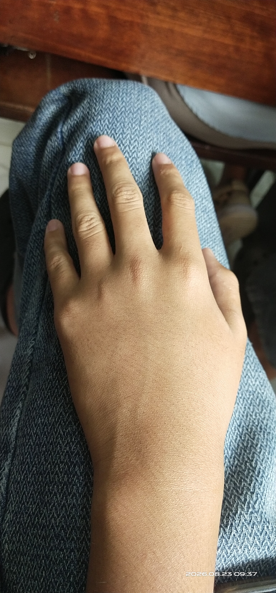 An overhead medium shot shows a person's left hand resting on their lap, which is covered by blue denim jeans. The hand is lightly tanned and positioned with the palm facing upwards. The fingers are slightly curled, with the thumb visible on the left side. The background consists of a wooden surface at the top and a blurry, out-of-focus area to the right, possibly indicating other people or objects. A timestamp, "2026.08.23 09:37," is visible in the lower right corner of the image. The scene is indoors, and there is no indication of the specific location within Jaar, Indonesia.