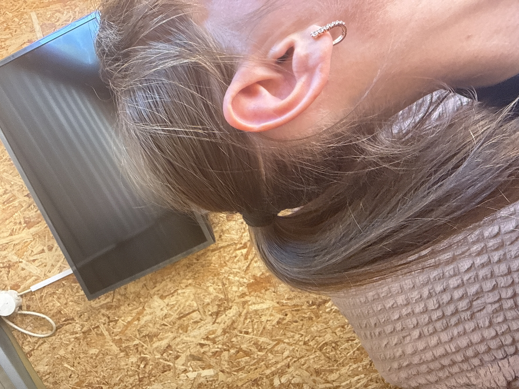 The image shows a close-up of a person's head and upper shoulder. The person has light brown hair pulled back into a low ponytail. The left ear is visible, adorned with a hoop earring containing small, clear stones along the upper edge. The person is wearing a light pink waffle-textured top. In the background, there is a dark-colored rectangular object mounted on a wall covered in light-colored, patterned wooden boards. A white cord with a white plug is also visible on the wooden wall, partially obscured by the rectangular object.