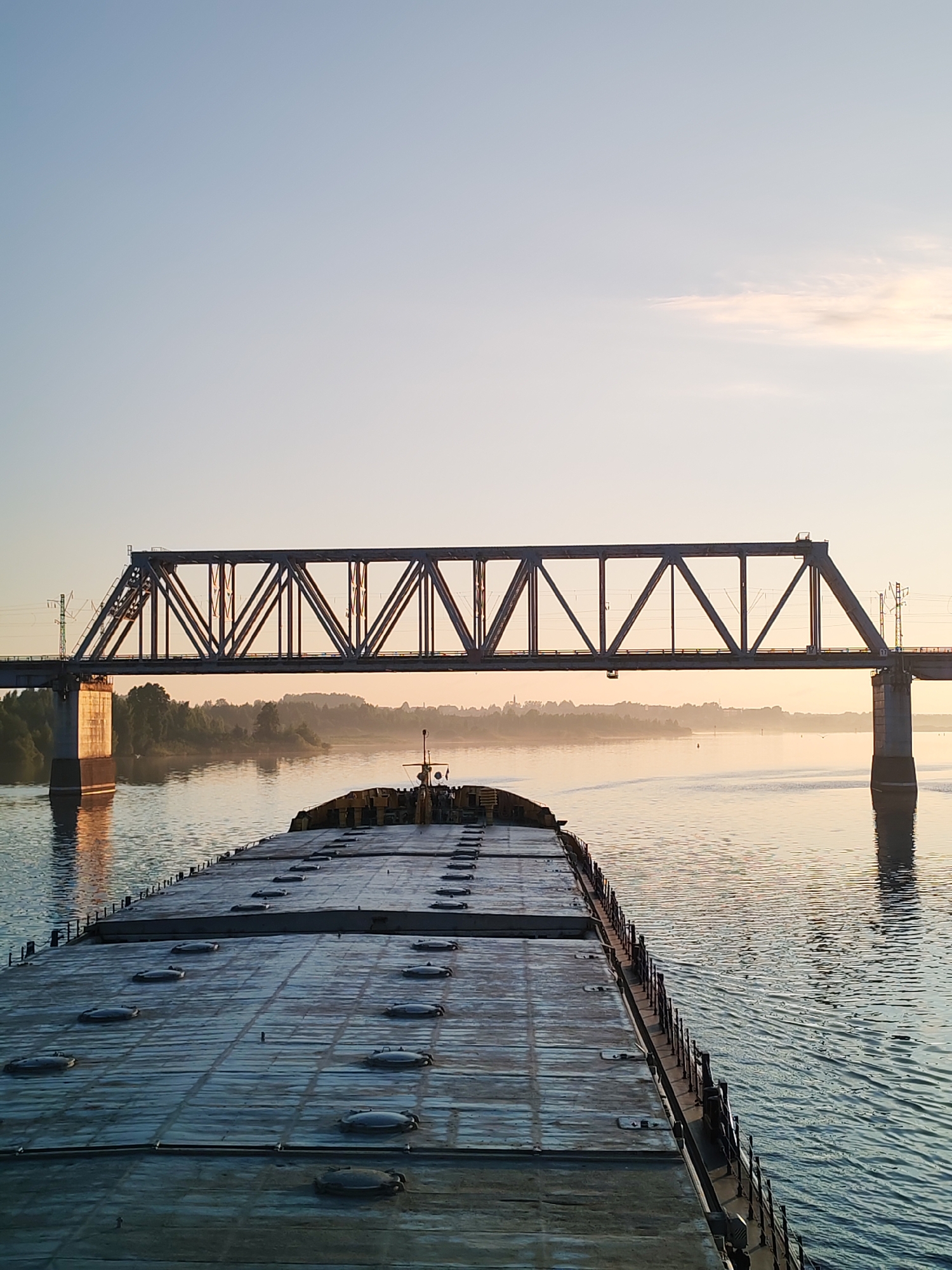 A barge filled with what appears to be hatch covers is traveling on a body of water with a truss bridge in the background. The bridge is a railroad bridge. The lighting suggests it is either sunrise or sunset. The location is likely the city of Нифантово, Russia.