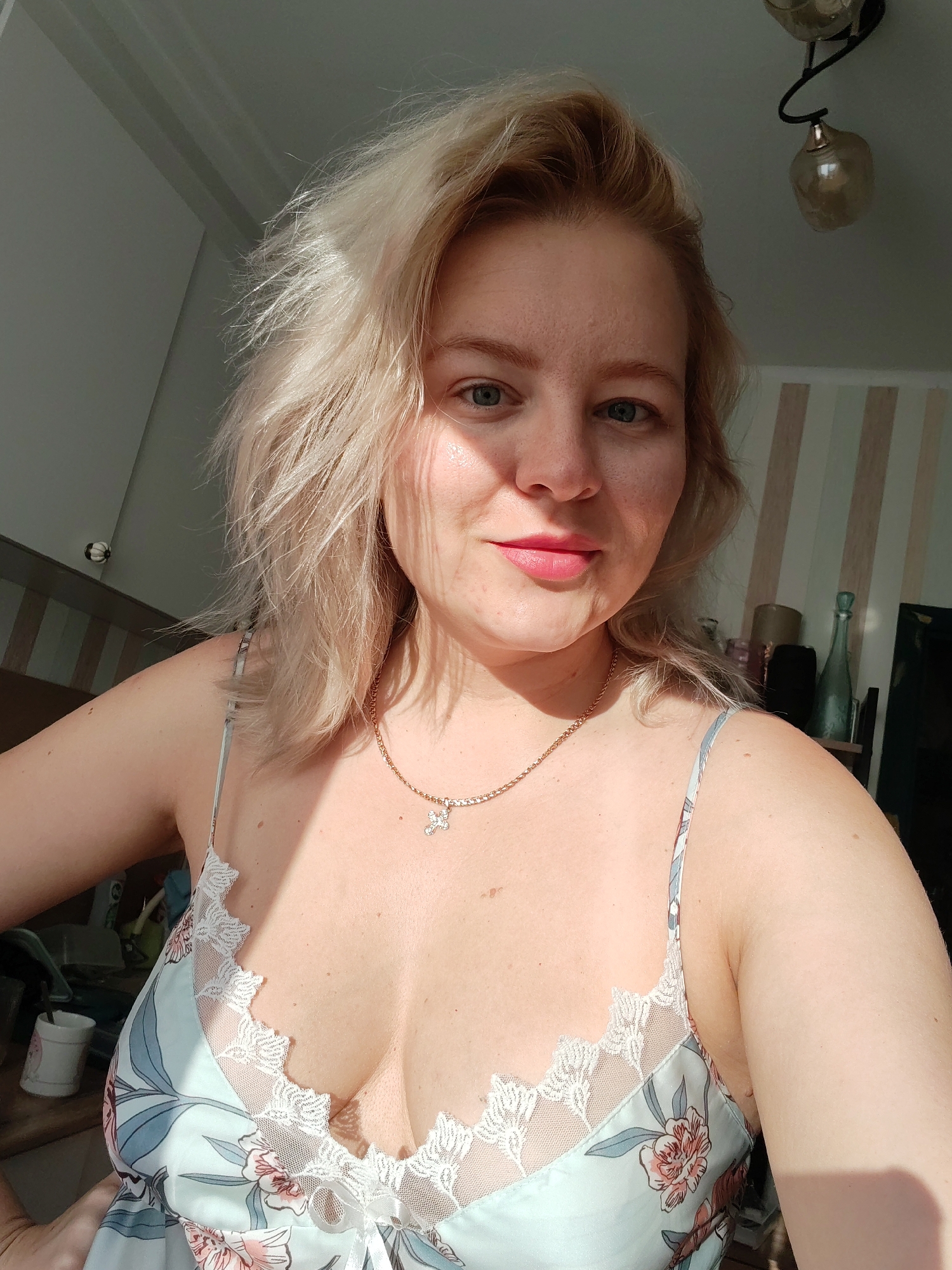 A woman with blonde hair and blue eyes is taking a selfie indoors. She is wearing a light blue floral print nightgown with white lace trim. She is also wearing a gold chain necklace with a cross pendant. The background shows a wall with vertical stripes and a shelf with various objects. The scene appears to be a domestic interior, potentially in the city of Izhevsk, Russia.