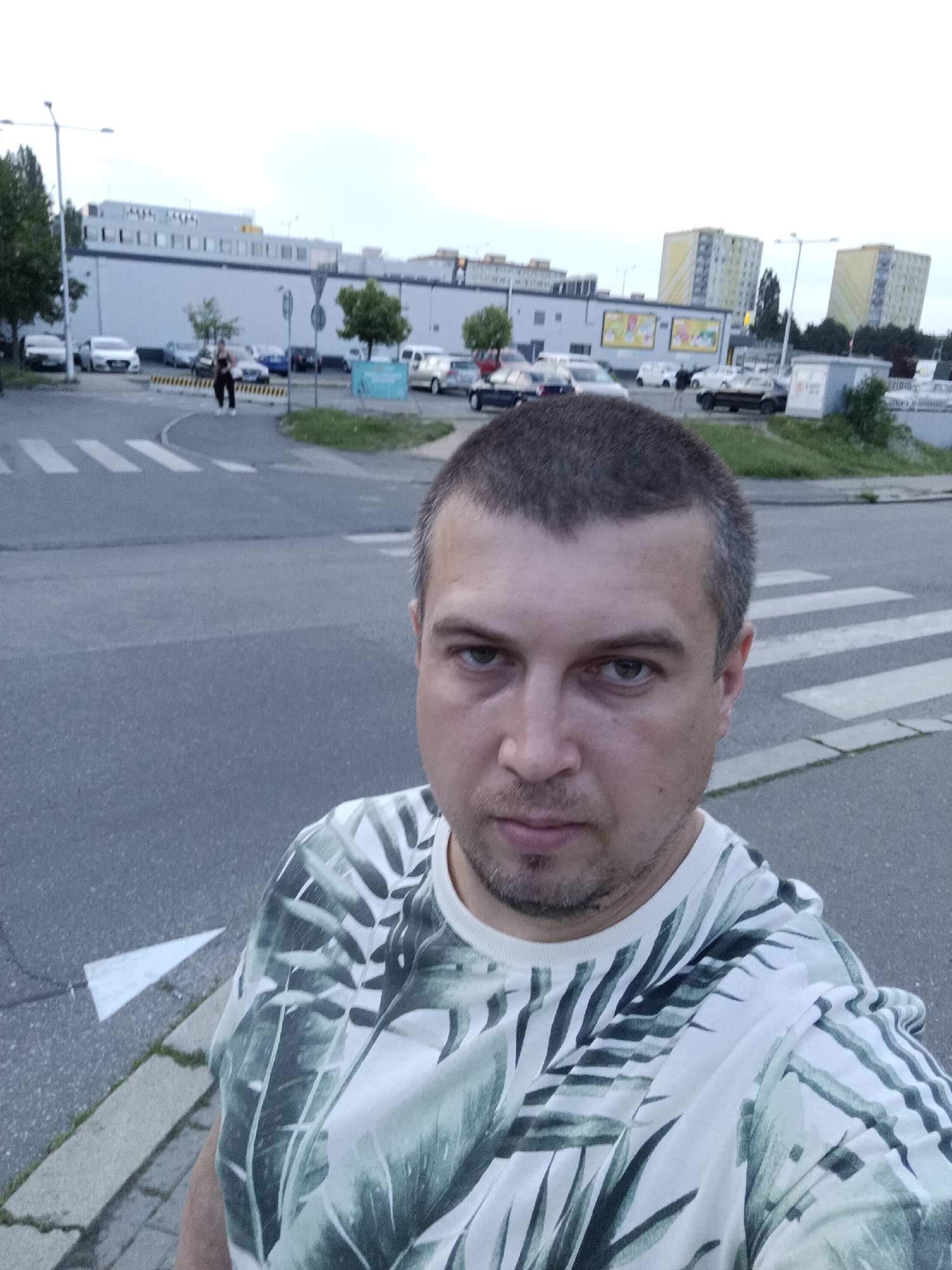 A man with short brown hair and a light beard takes a selfie outdoors. He is wearing a white t-shirt with a tropical leaf pattern in green and grey. The background shows a paved area with a pedestrian crossing, a street with several parked cars, a building with a white facade and a sign, and two yellow apartment buildings in the distance. The scene appears to be in a suburban area of Prague, Czechia. The sky is overcast.
